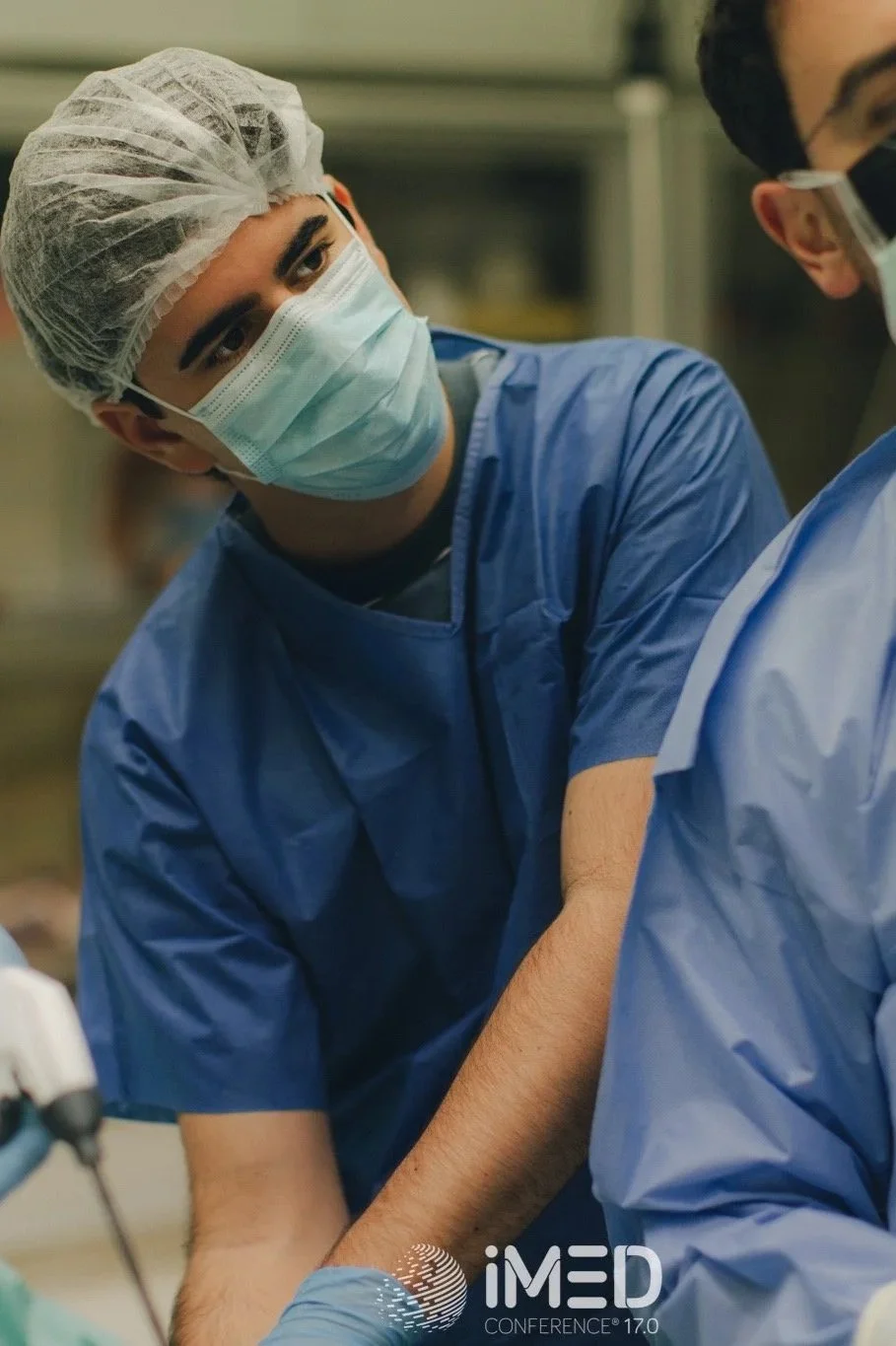 A young male doctor or surgeon in scrubs, face mask, and hair cover, assisting in a surgical procedure while intently observing, in a professional medical setting.