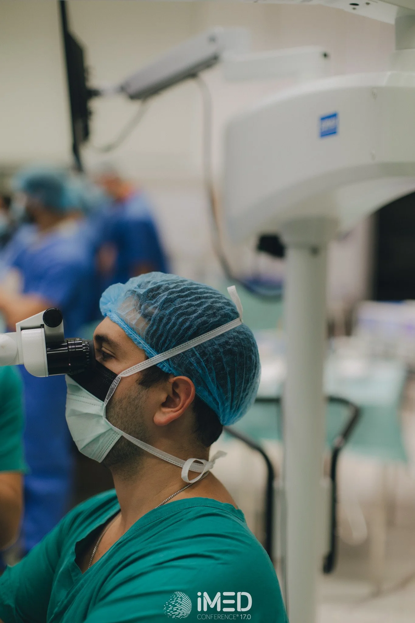 A male surgeon in scrubs, surgical mask, and cap operating with a microscope in a medical environment, with other medical staff in the background.