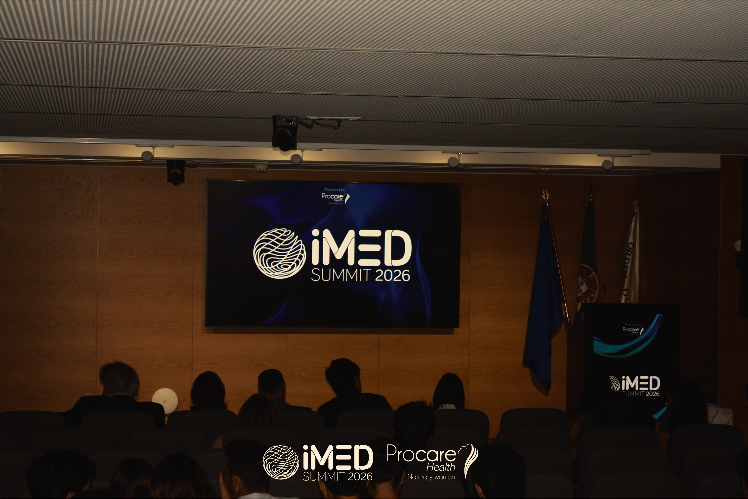 Conference room with audience watching a presentation on a large screen about the iMED Conference 17.0, with blue lighting and aurora borealis imagery on the screen.