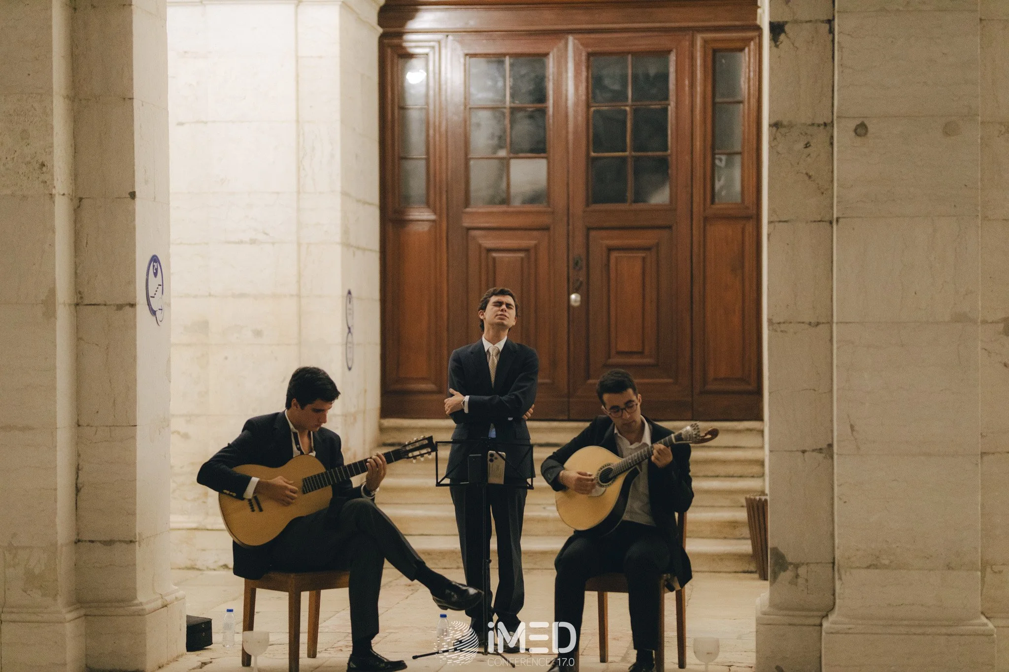 Three men in suits performing classical guitar and singing in an elegant, grand hall with large wooden doors in the background.