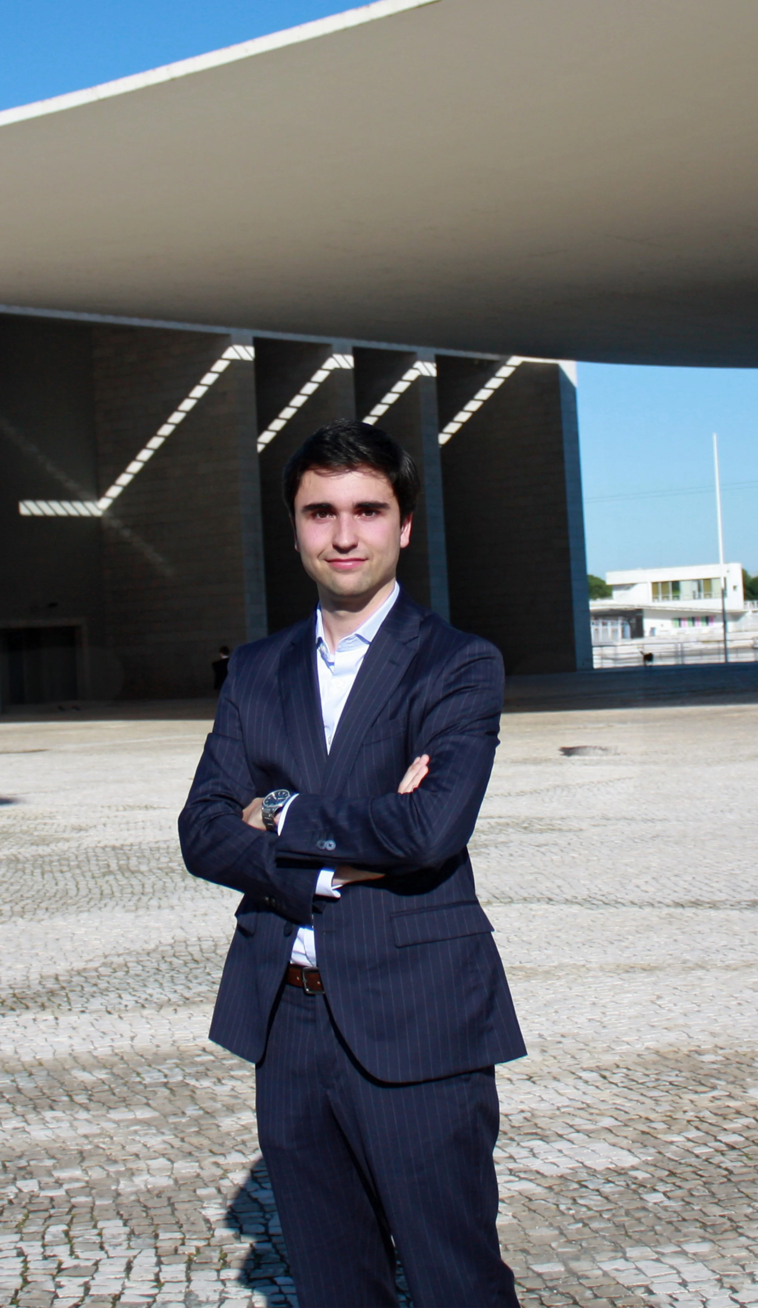 A young man in a dark pinstripe suit with crossed arms standing outside on a cobblestone pavement under a modern building with geometric design.