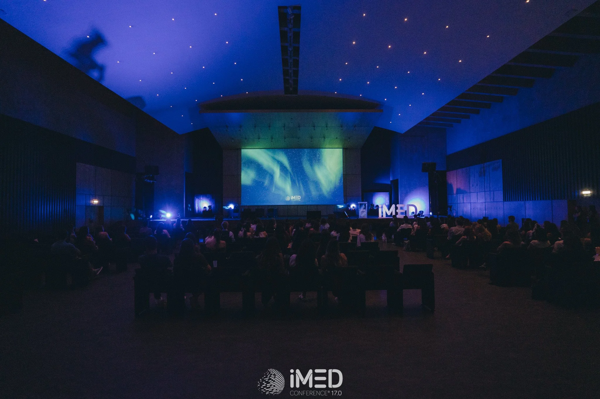 Conference room with audience watching a presentation on a large screen about the iMED Conference 17.0, with blue lighting and aurora borealis imagery on the screen.