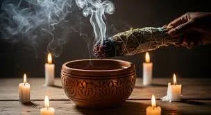 A hand holding a burning bundle of sage above a decorated bowl surrounded by lit candles.