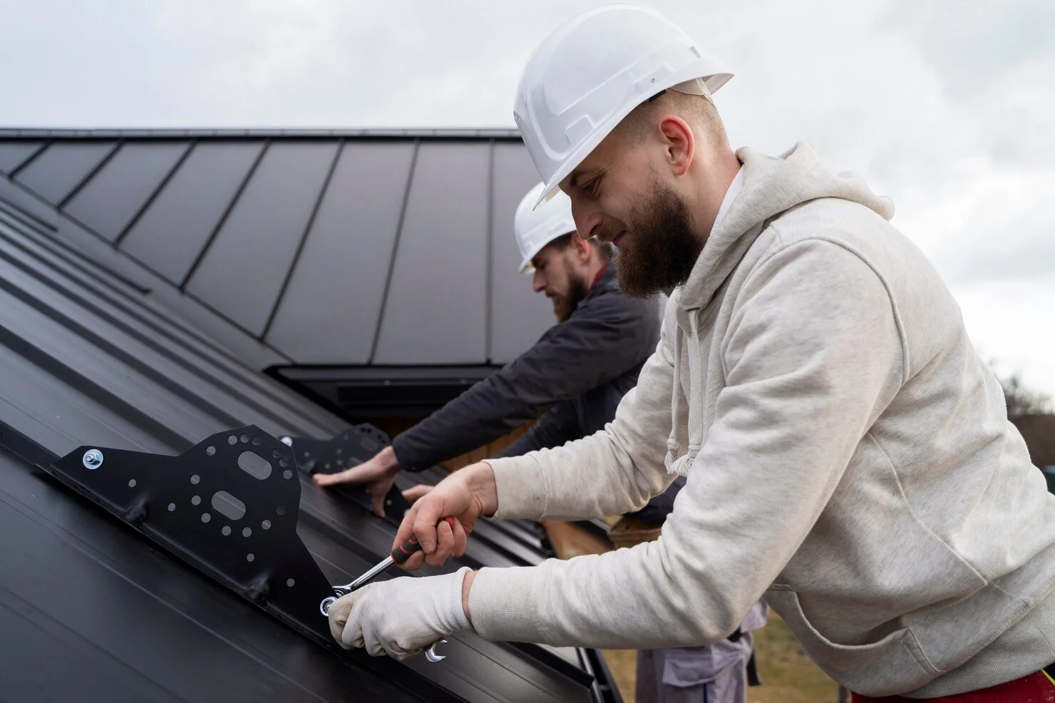 Worker with a laptop standing on the edge of a brick rooftop, while another worker kneels on a white flat roof holding a board.