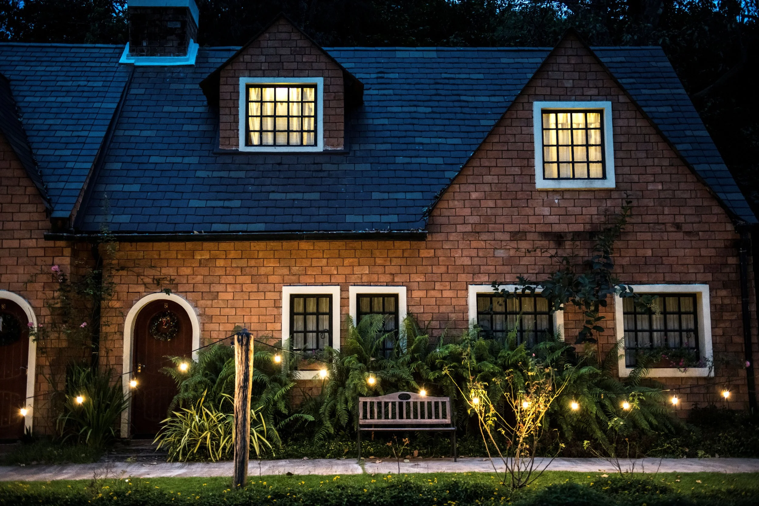 A brick house with a dark shingled roof, illuminated windows, and holiday wreaths on the front doors. There are string lights hanging in the garden area in front of the house, along with a wooden bench and some plants and bushes.