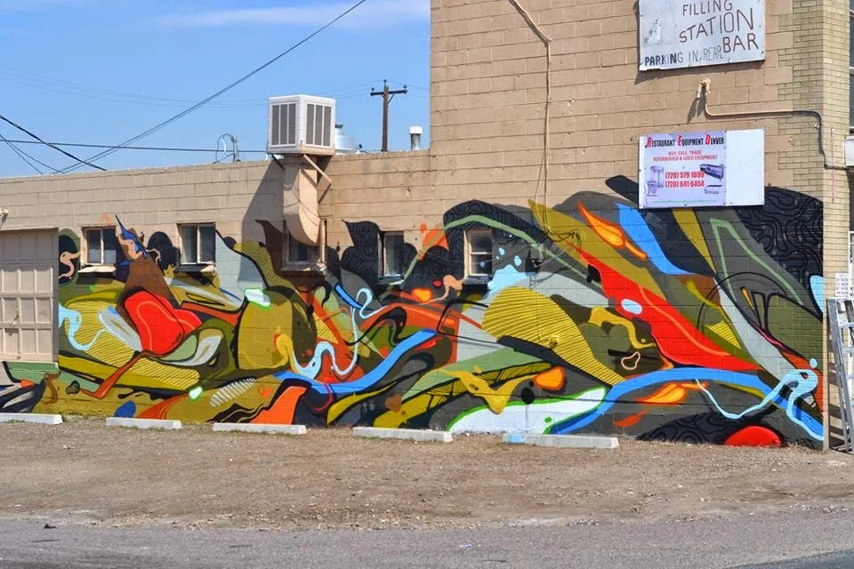 Colorful abstract graffiti mural on a brick wall with small windows, located next to signage for a filling station, parking, and bar.