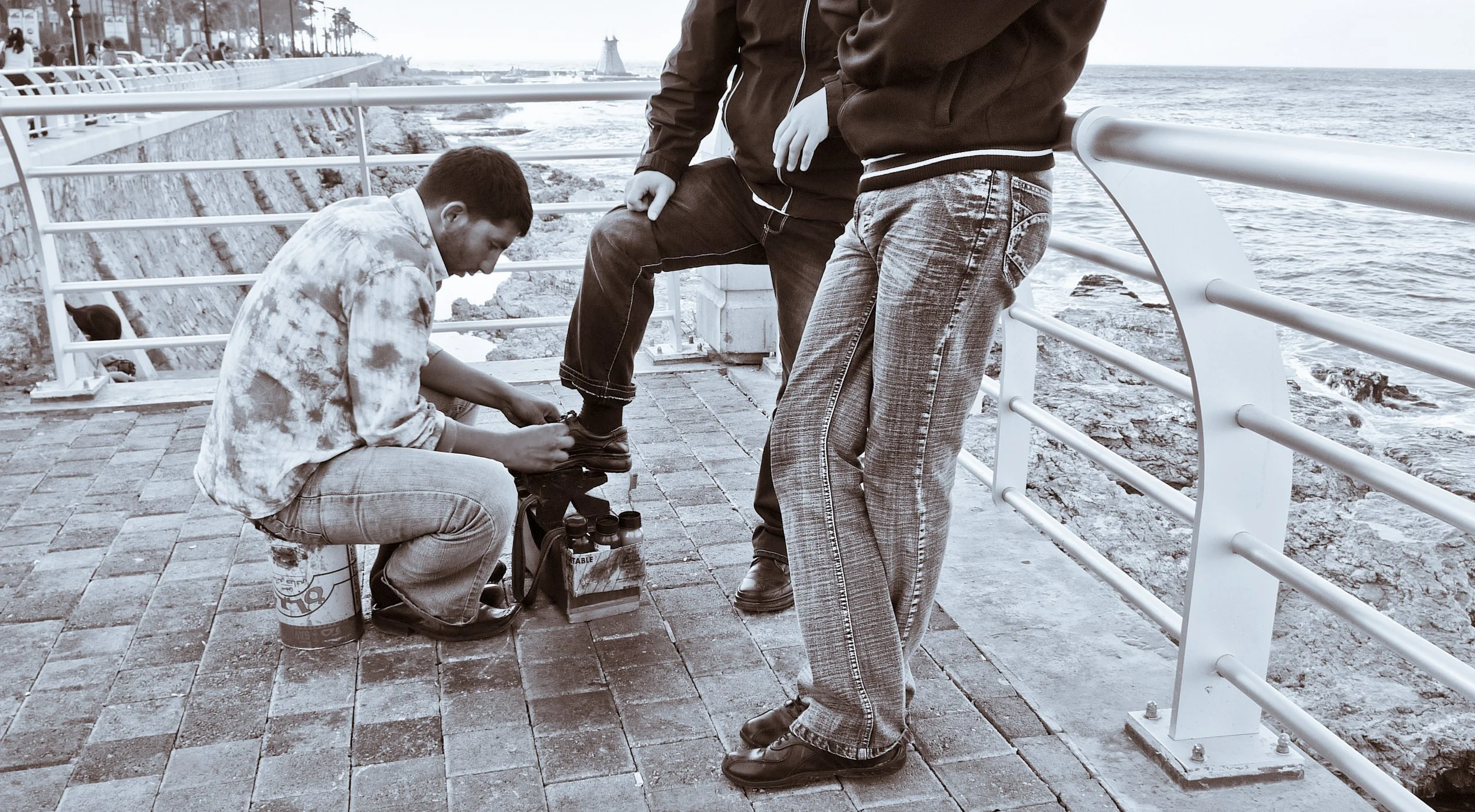 A man kneeling on a brick walkway by the ocean helps another man tie his shoelaces. The scene is in black and white and shows a railing along the waterfront.