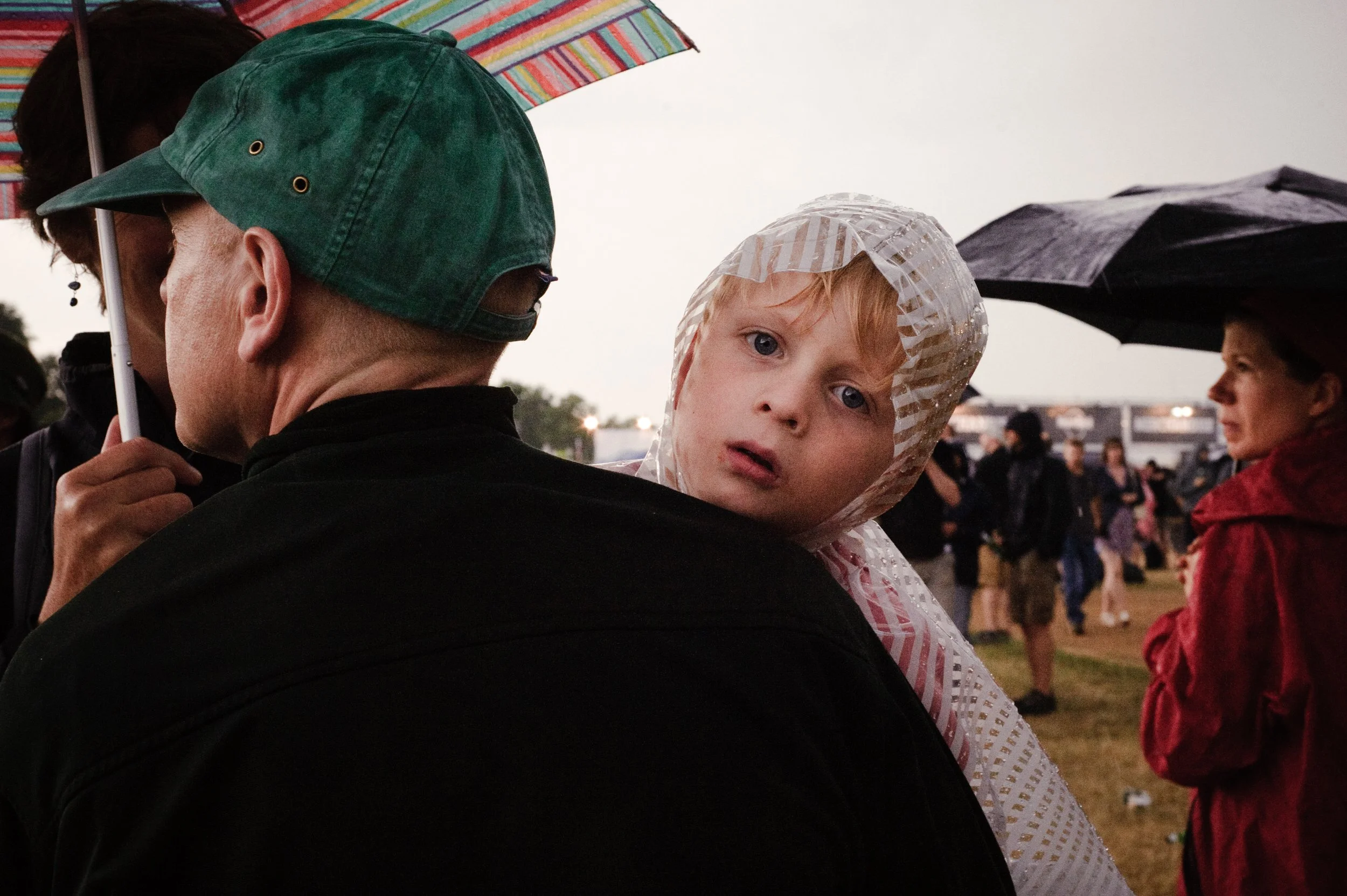 A young child with blond hair and blue eyes, wearing a transparent rain poncho, is resting on an adult's shoulder during a rainy outdoor event, with other people holding umbrellas in the background.