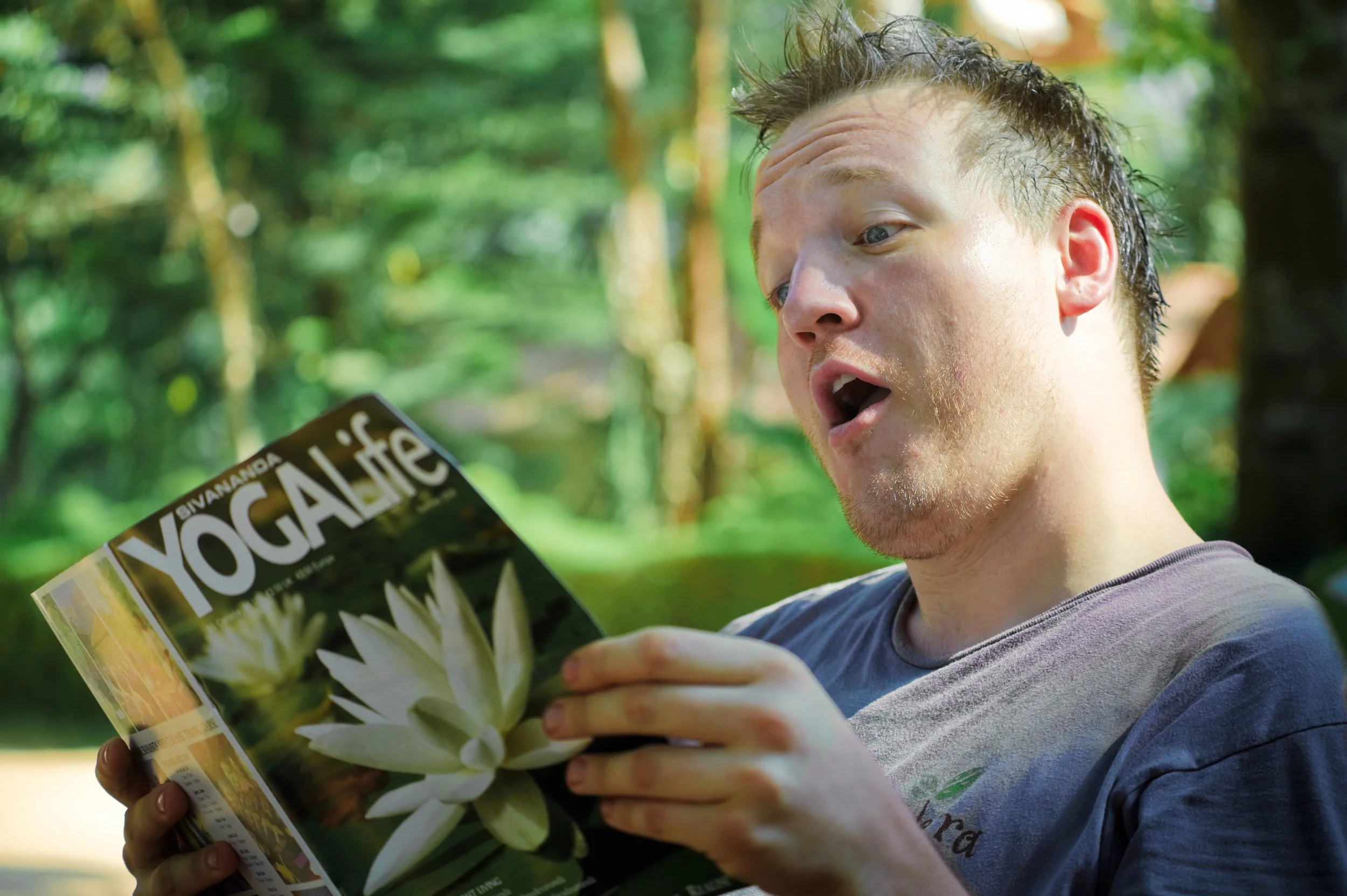 A man in an outdoor, green, wooded area looks surprised as he reads a magazine titled "YOGA GALIC" featuring a close-up of a white flower on the cover.