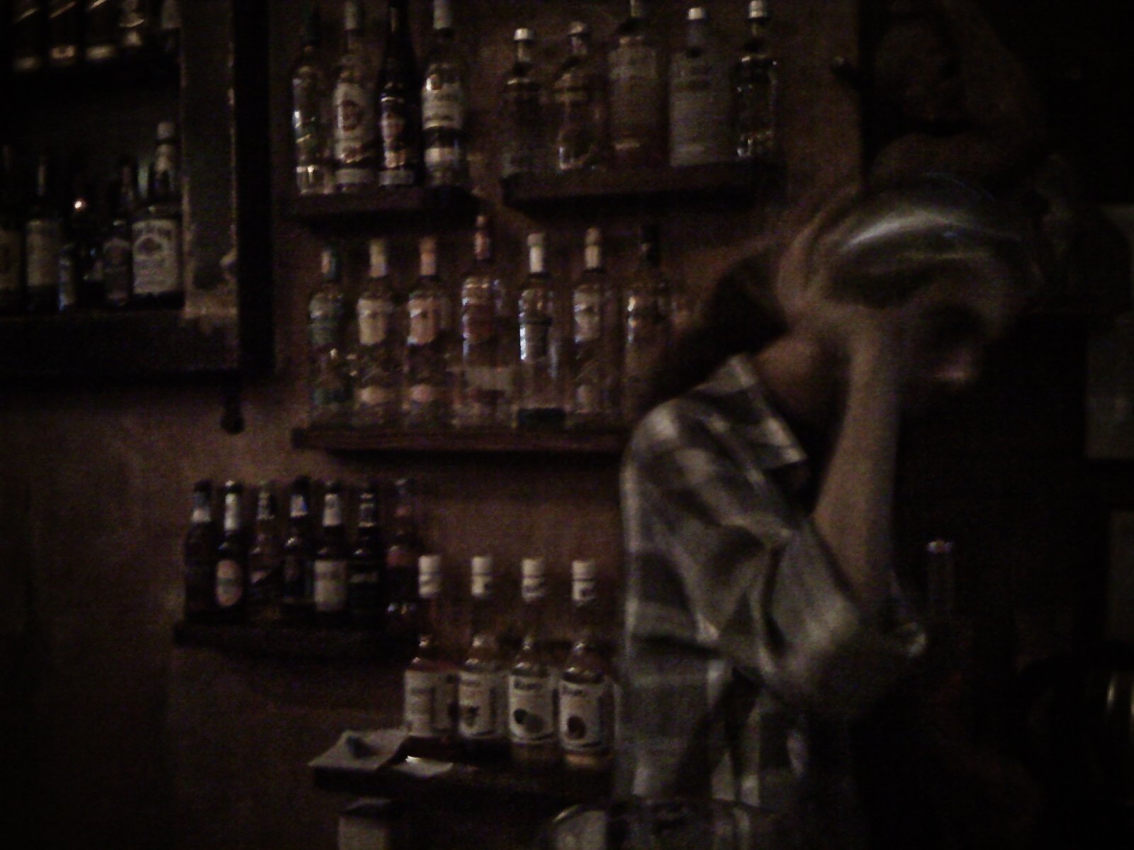 A woman wearing a plaid shirt and a helmet, standing in a dimly lit bar with shelves of liquor bottles behind her.