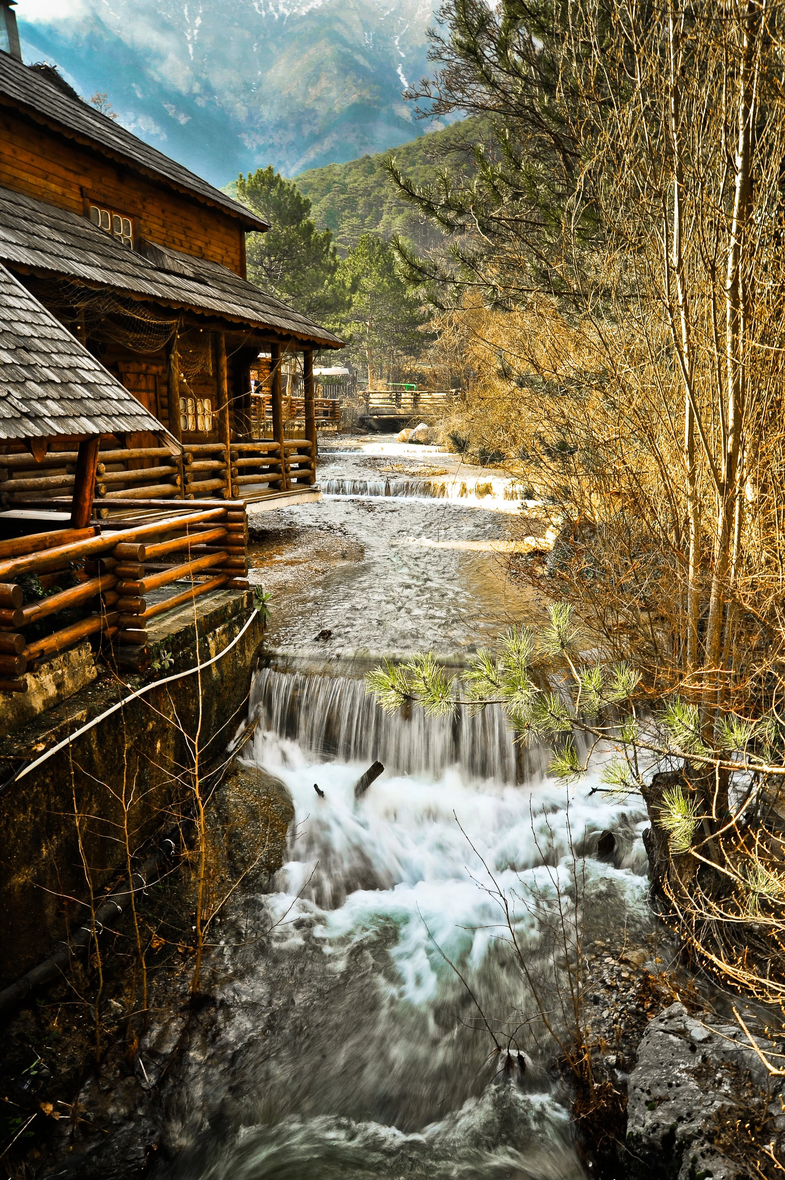 A rustic wooden cabin with a shingled roof situated by a flowing creek surrounded by trees and mountains in the background.