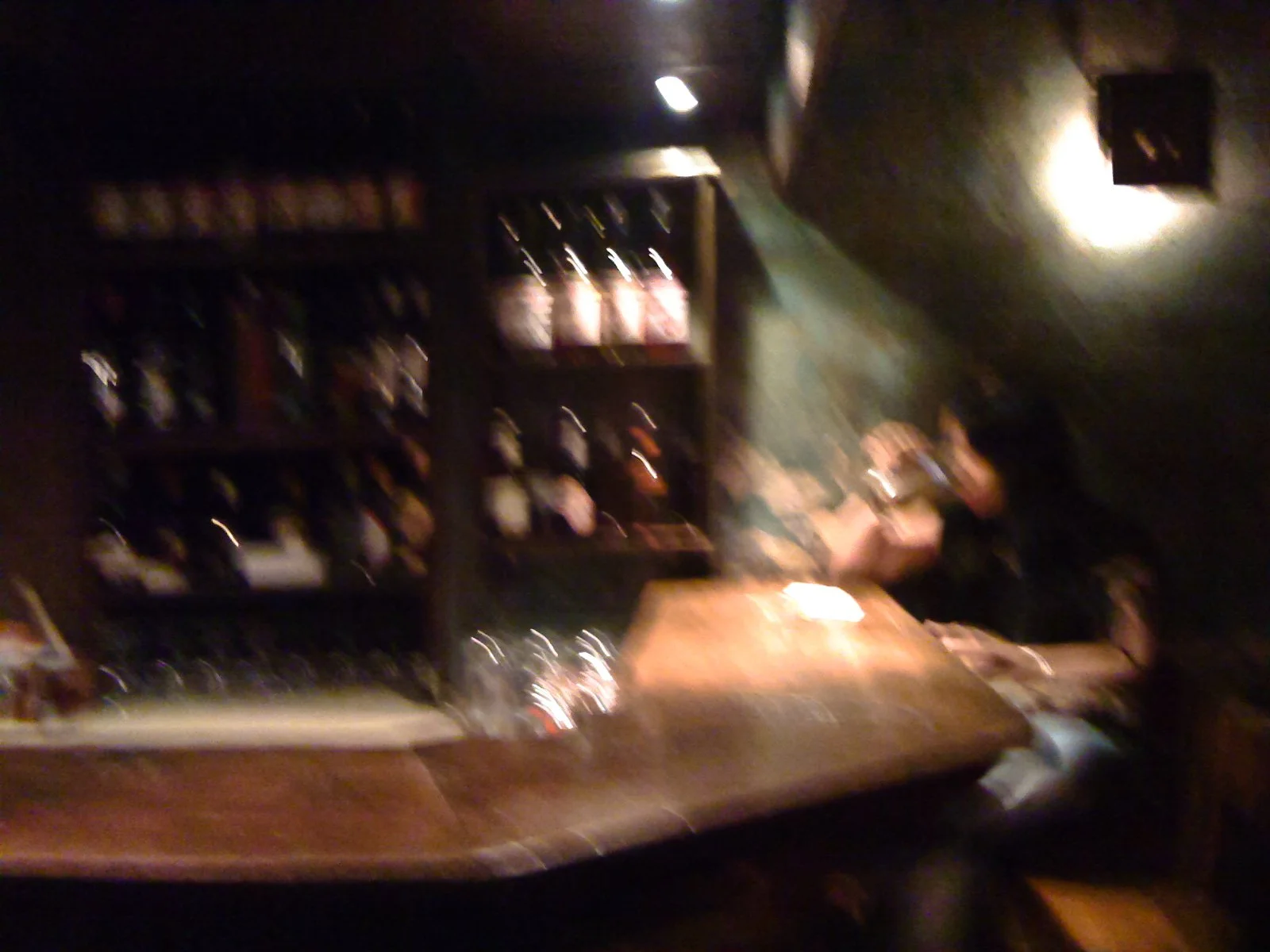 Blurred image of a bartender preparing drinks behind a wooden bar counter with shelves of liquor bottles in a dimly lit bar or pub.