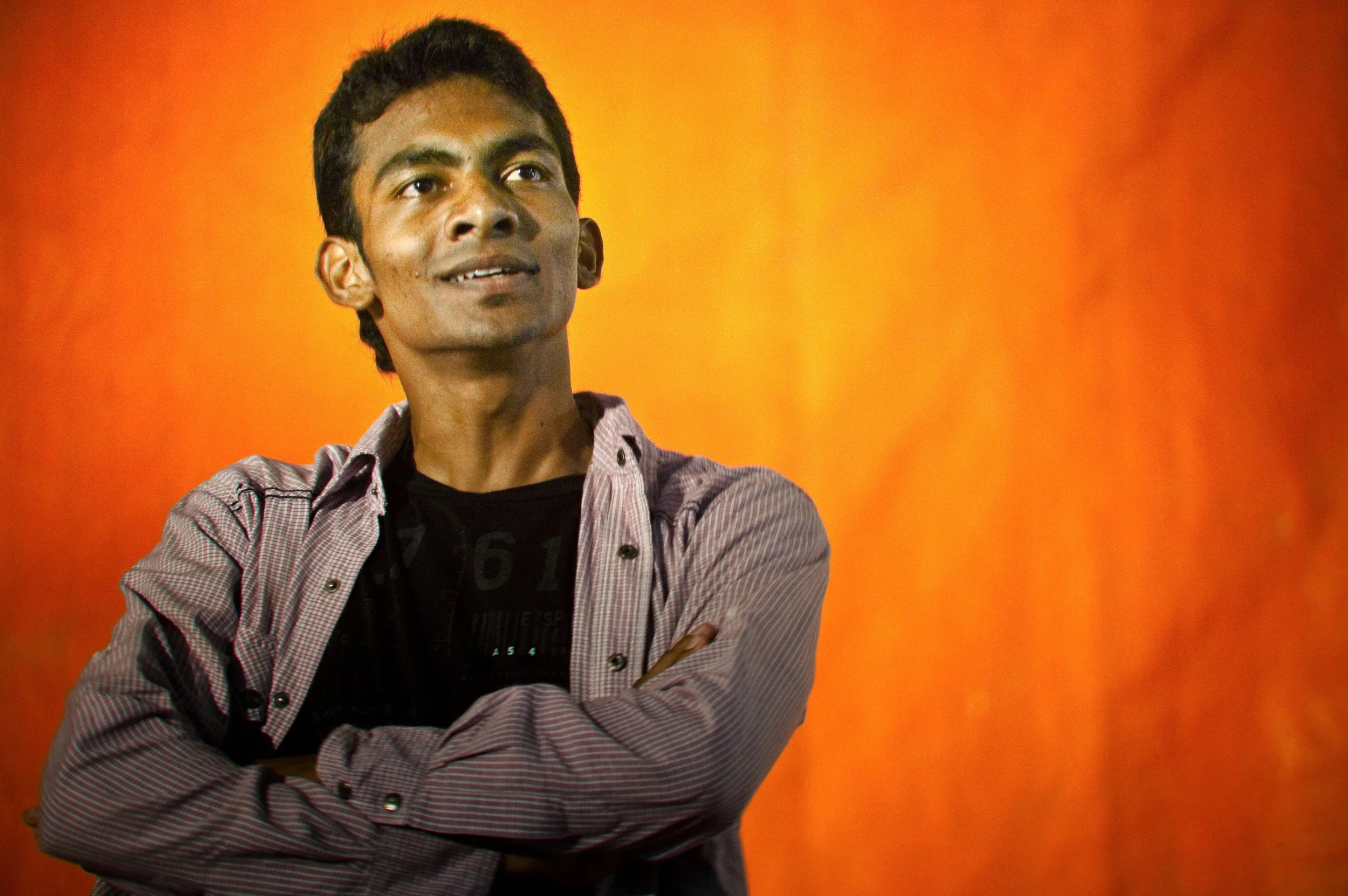 A young man with dark hair and brown skin standing with his arms crossed, looking slightly upward, against a vibrant orange background.