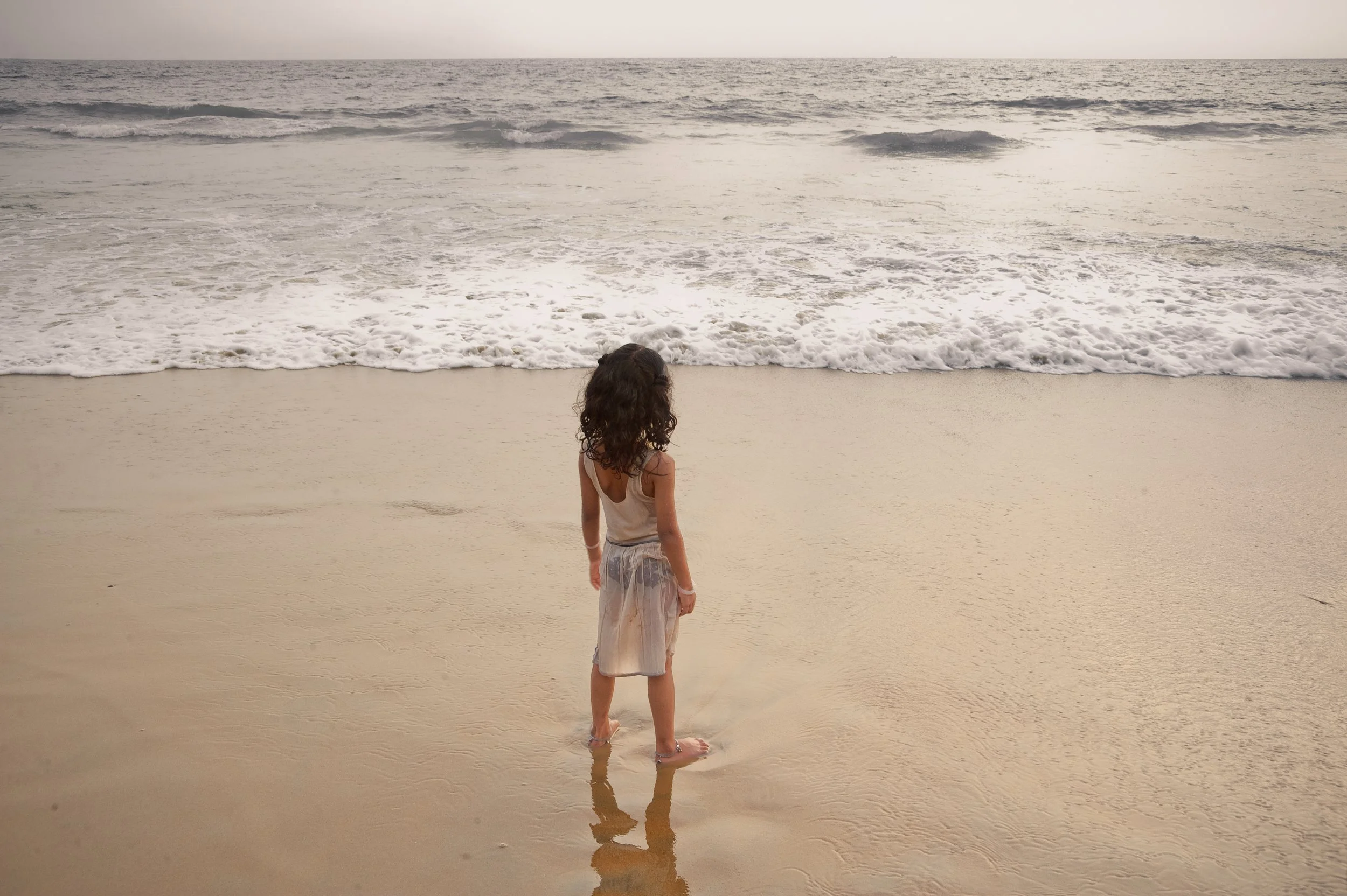 A young girl with curly hair wearing a sleeveless top and a sheer skirt standing at the shoreline of a beach, looking at the ocean waves.