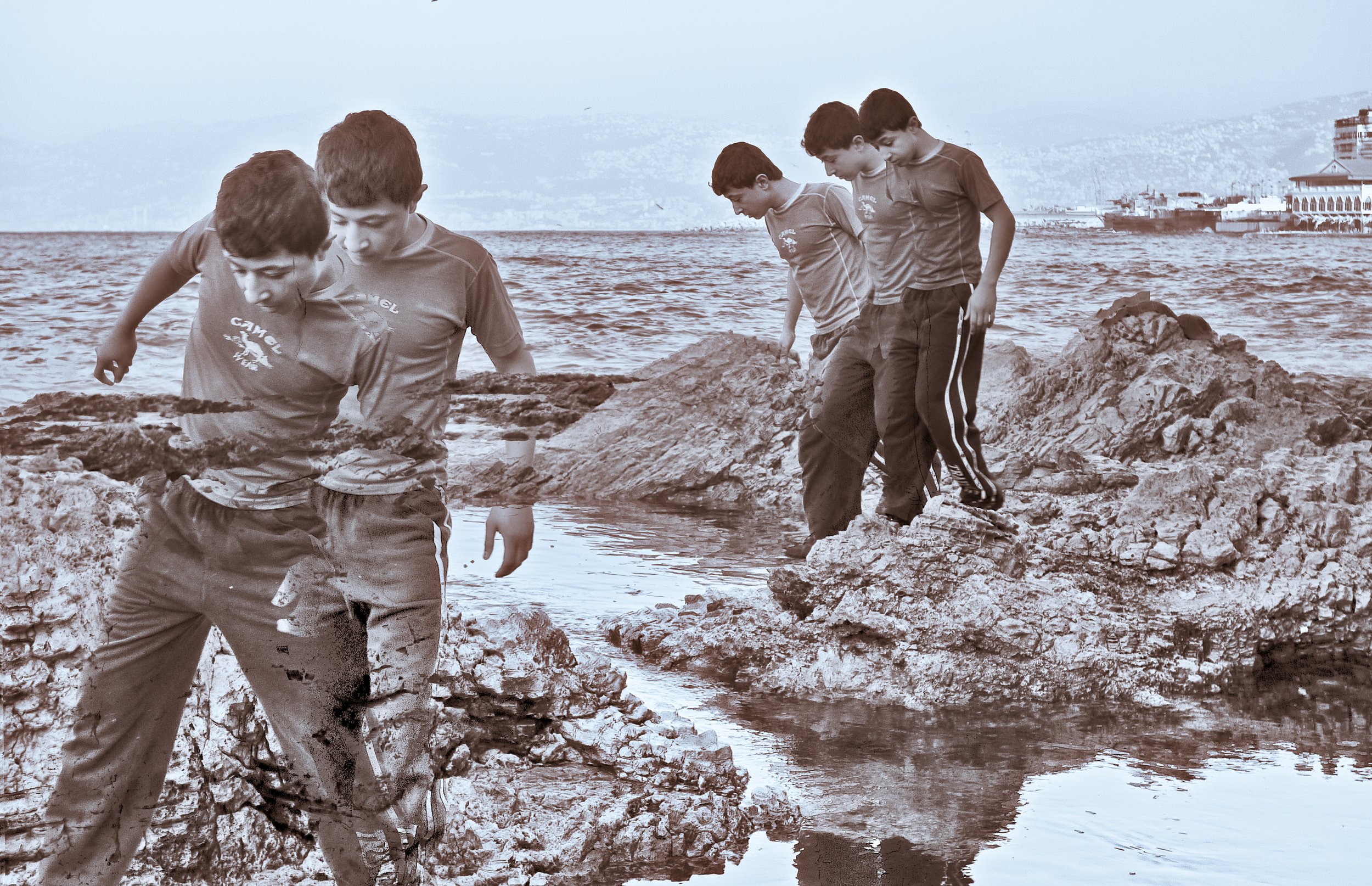 Multiple boys standing and walking on rocks near a body of water by the coast, with buildings and hills in the background.