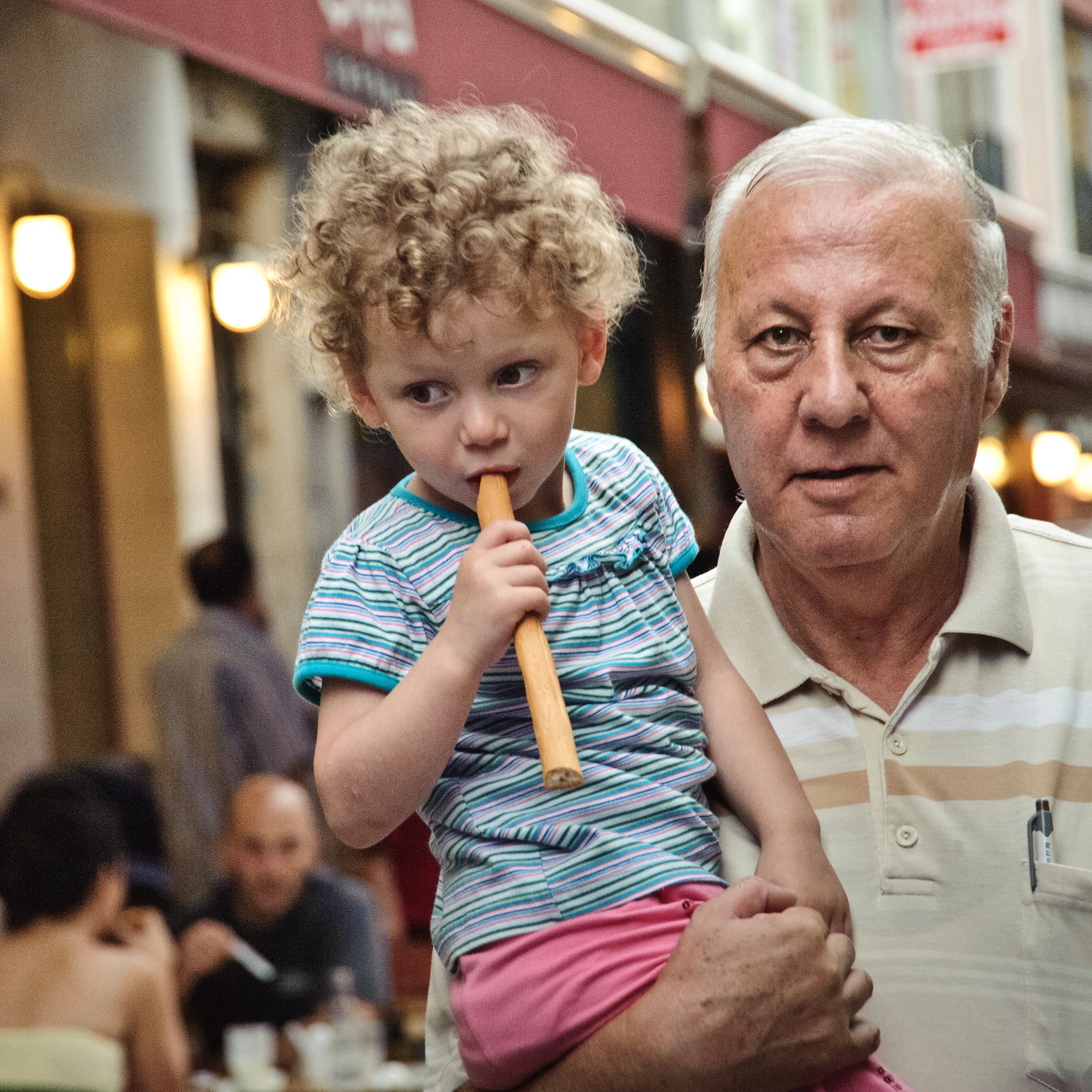 An elderly man holding a young girl with curly hair in a busy, well-lit indoor space. The girl is holding a stick-like object near her mouth and appear to be looking to the side.