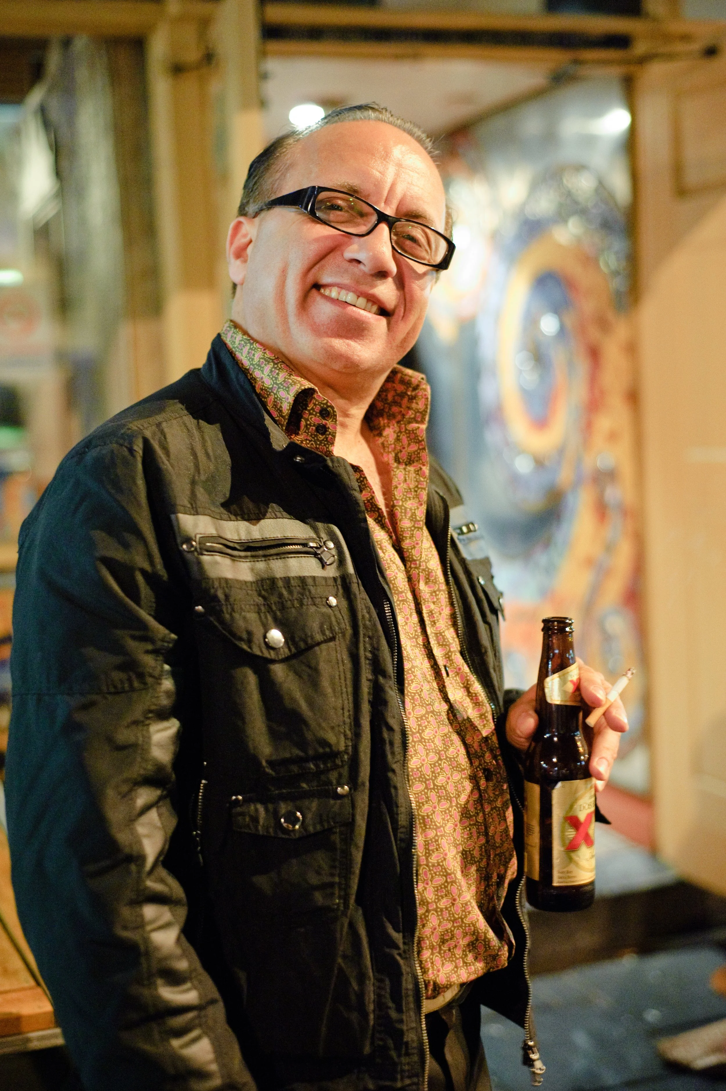 A middle-aged man with glasses and slicked-back hair smiling while holding a beer bottle and a cigarette in a casual indoor setting.
