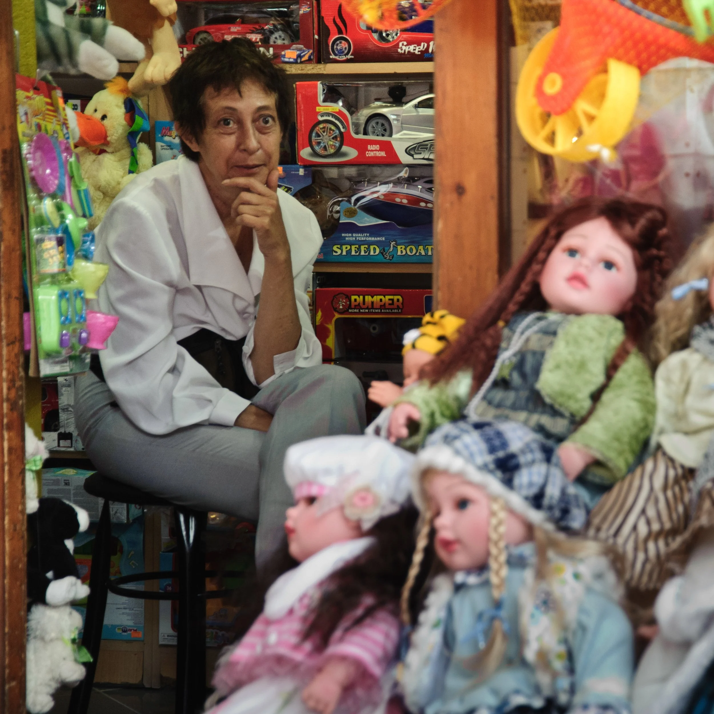 A woman with short dark hair, wearing a white shirt, sitting in a toy store surrounded by dolls and stuffed animals. She is looking at the camera with a curious expression, resting her chin on her hand. The background is filled with toy cars and boxe
