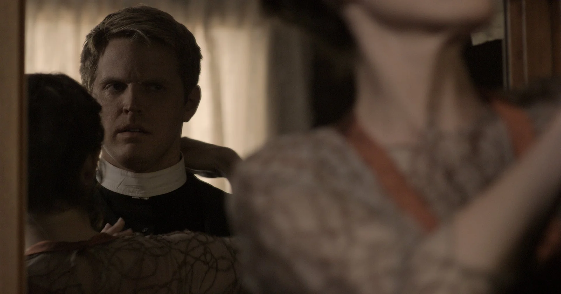 A priest looks into a mirror partially obscured by a woman and another person in the foreground.