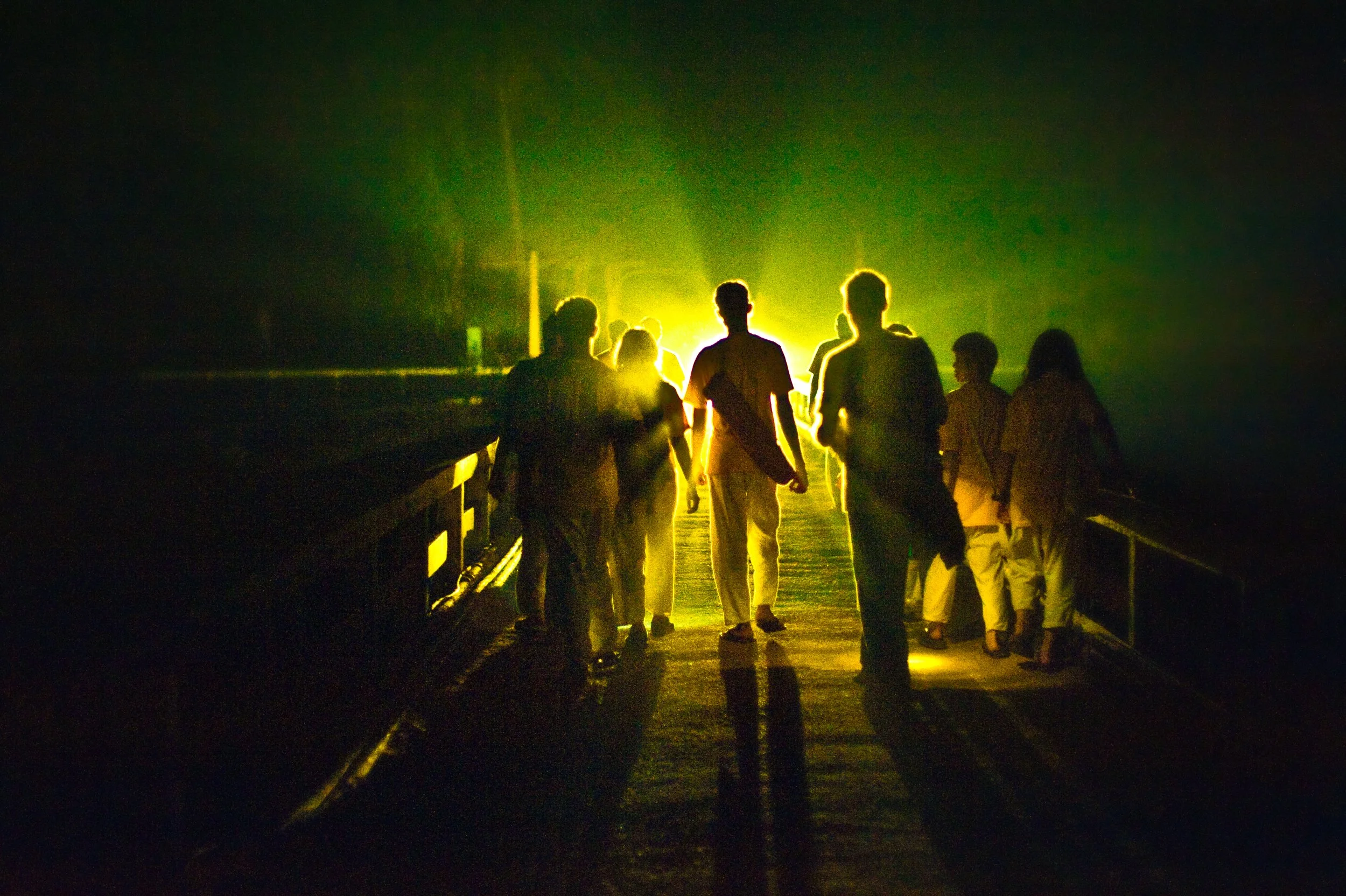 Group of people walking on a bridge at night with bright light in the background.