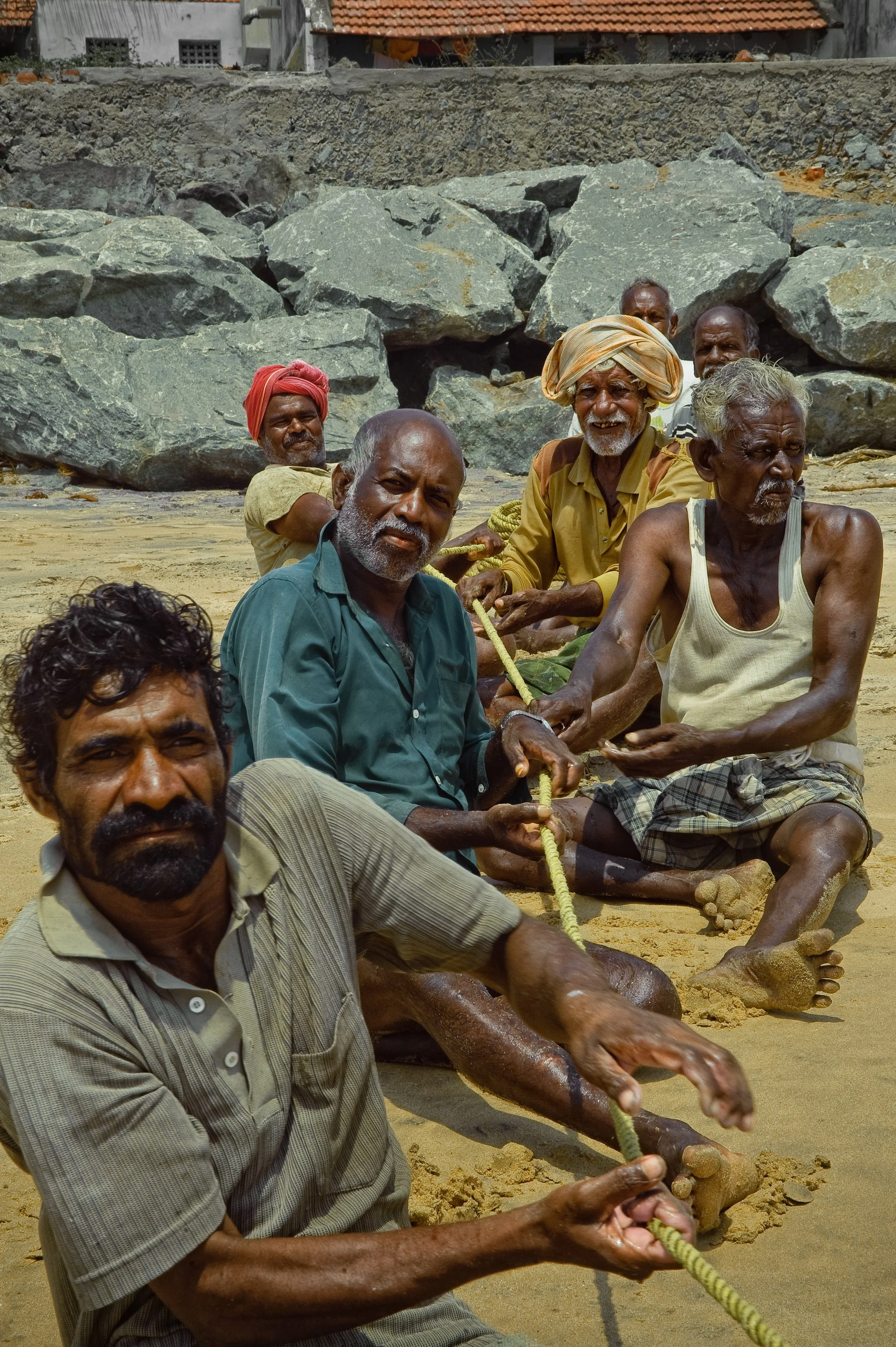 Group of men sitting on a sandy beach, holding a rope together, with rocky formations and buildings in the background.
