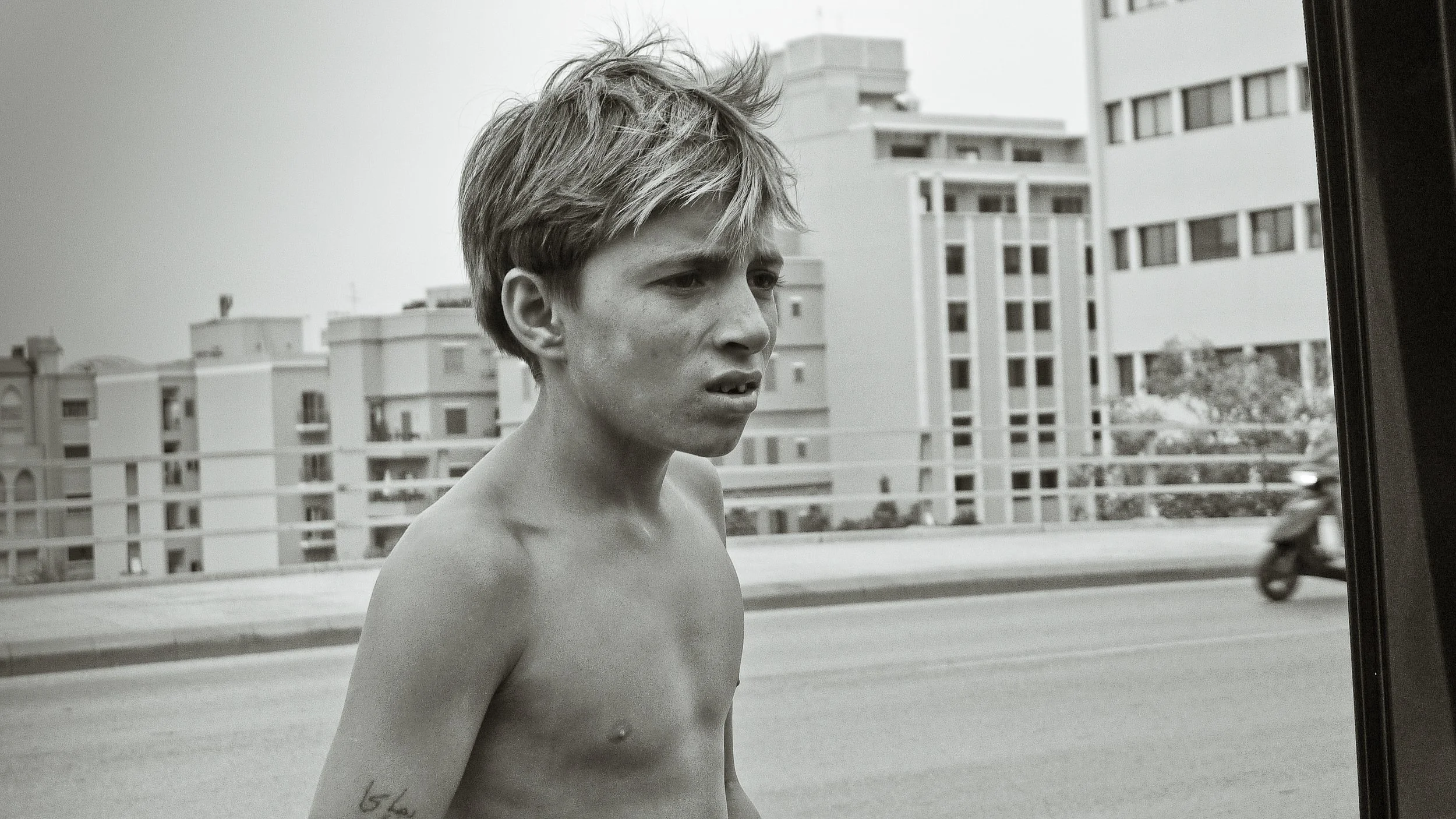 A shirtless young man with messy hair standing on a city street, looking off to the side with a concerned expression. Buildings and a blurred scooter are visible in the background.