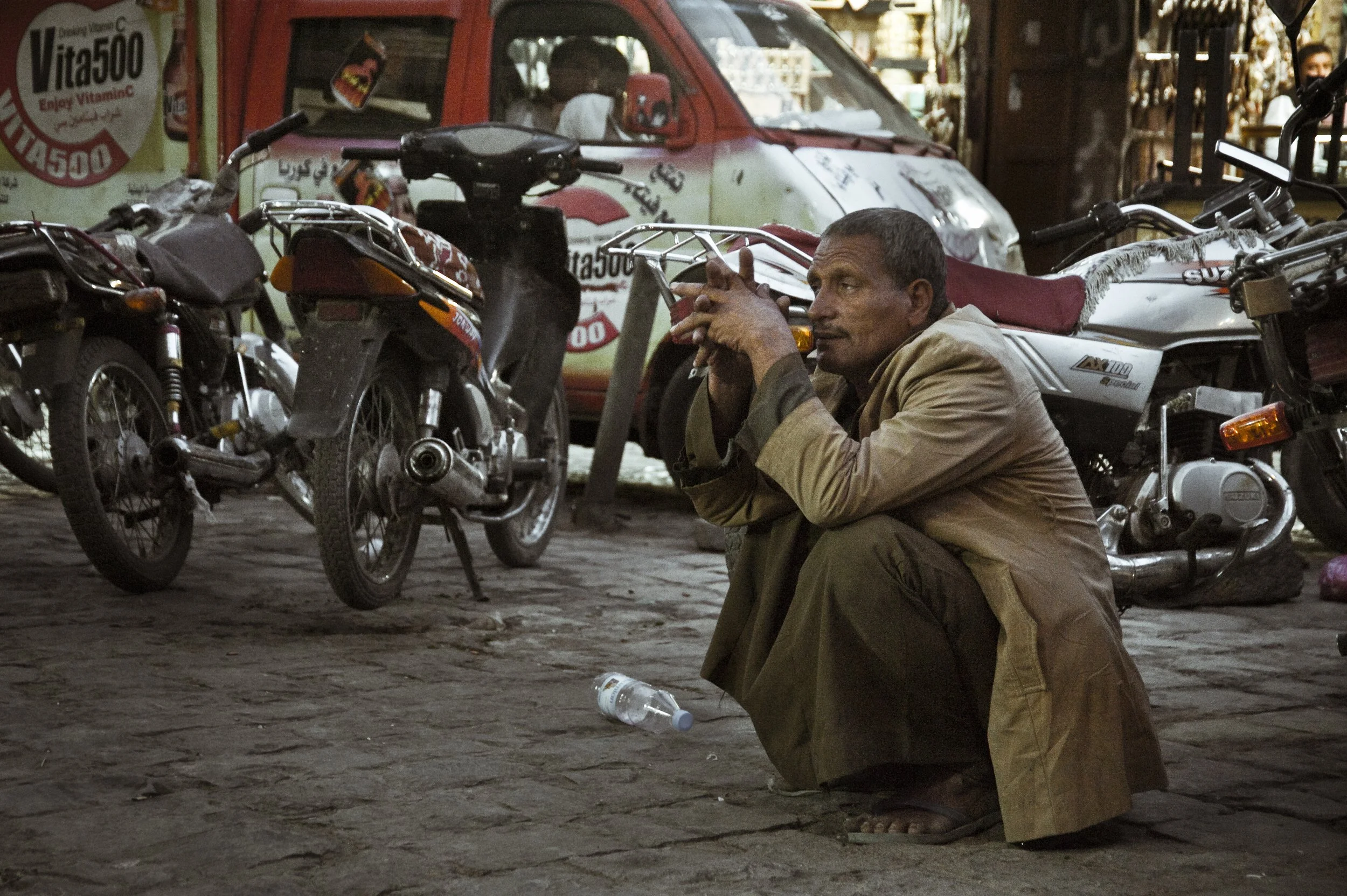 A man in a beige coat squats on a cobblestone street near parked motorcycles, with a red and white vehicle in the background, and a discarded plastic bottle on the ground.