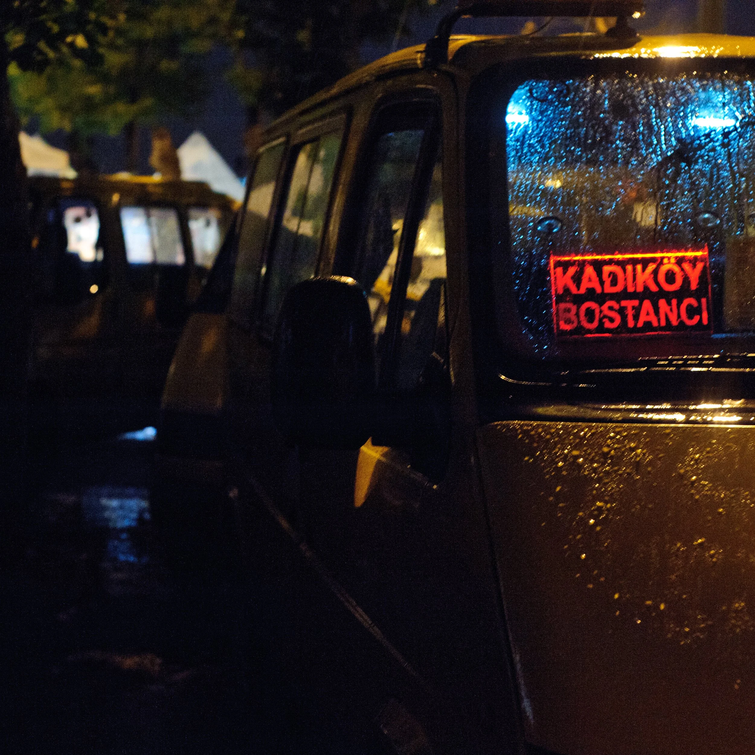 A close-up of a rain-covered vehicle with a red illuminated sign in the window that reads 'KADIKÖY BOSTANCI' in Turkish, taken at night.