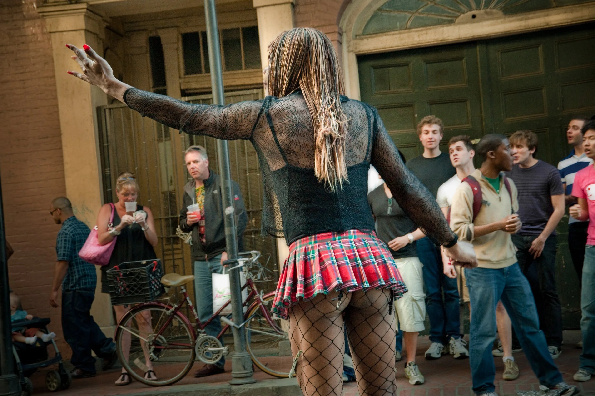 A woman dressed in black fishnet clothing with a red plaid skirt and blonde hair dancing on a city street, surrounded by a crowd of people including a woman with a pink bag and people taking photos.