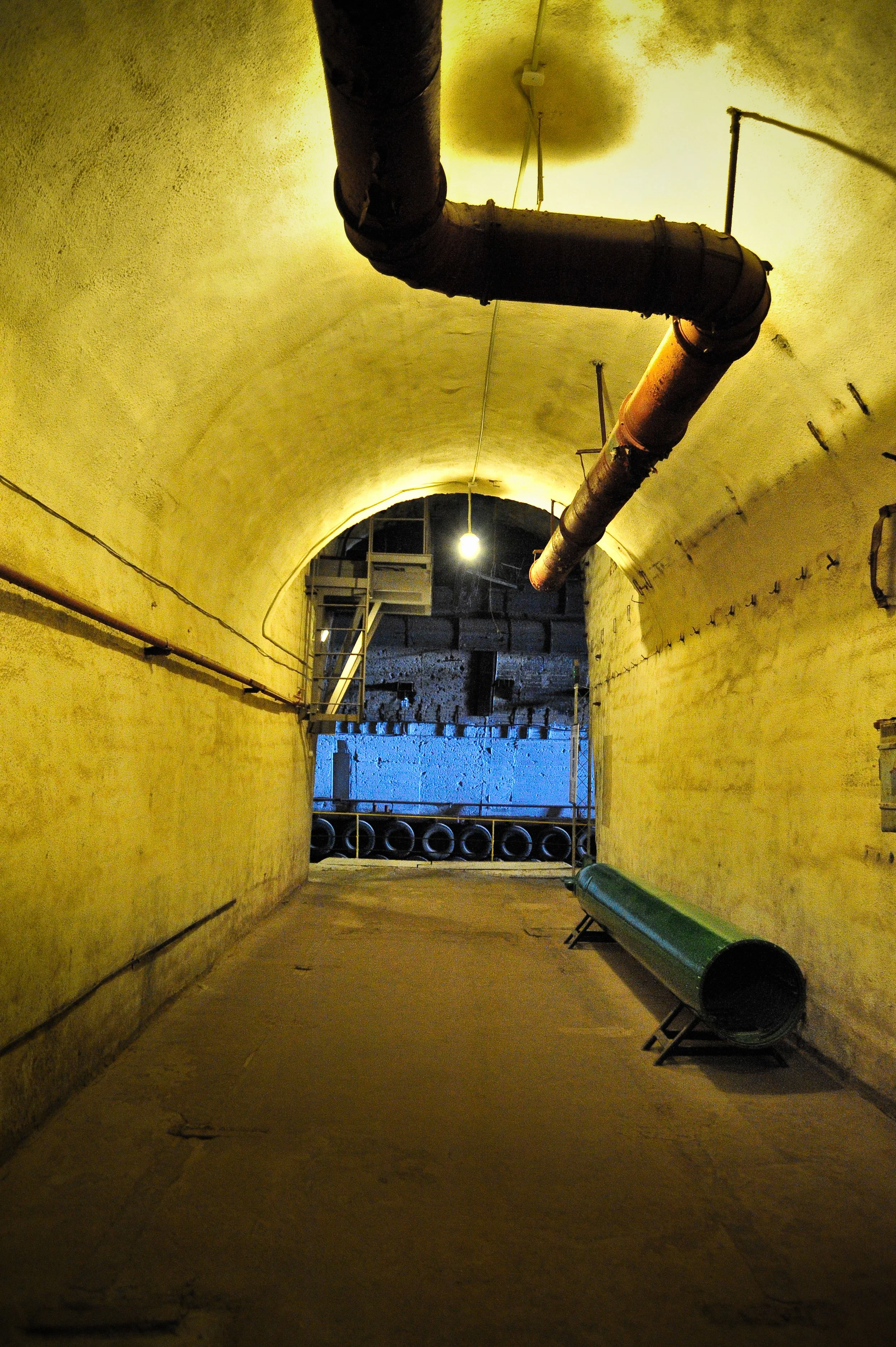Underground tunnel with yellowish walls, large pipes hanging from the ceiling, and a bench on the right side, illuminated by a single light bulb.