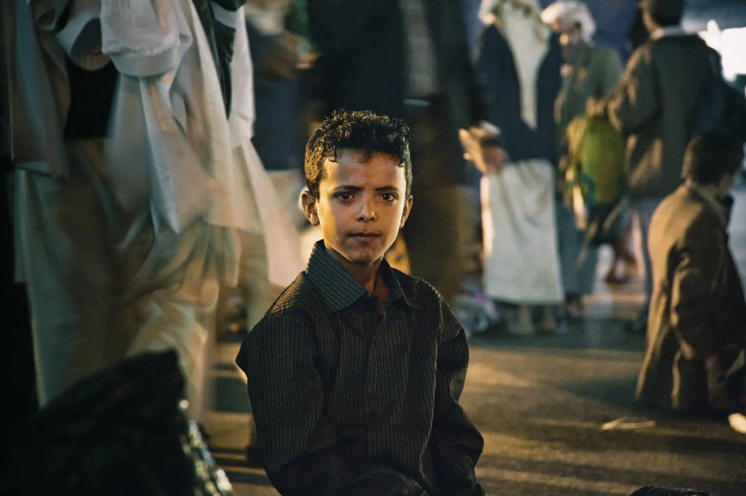 A young boy sitting outdoors in a busy market or gathering at night, surrounded by people in traditional and casual clothing.