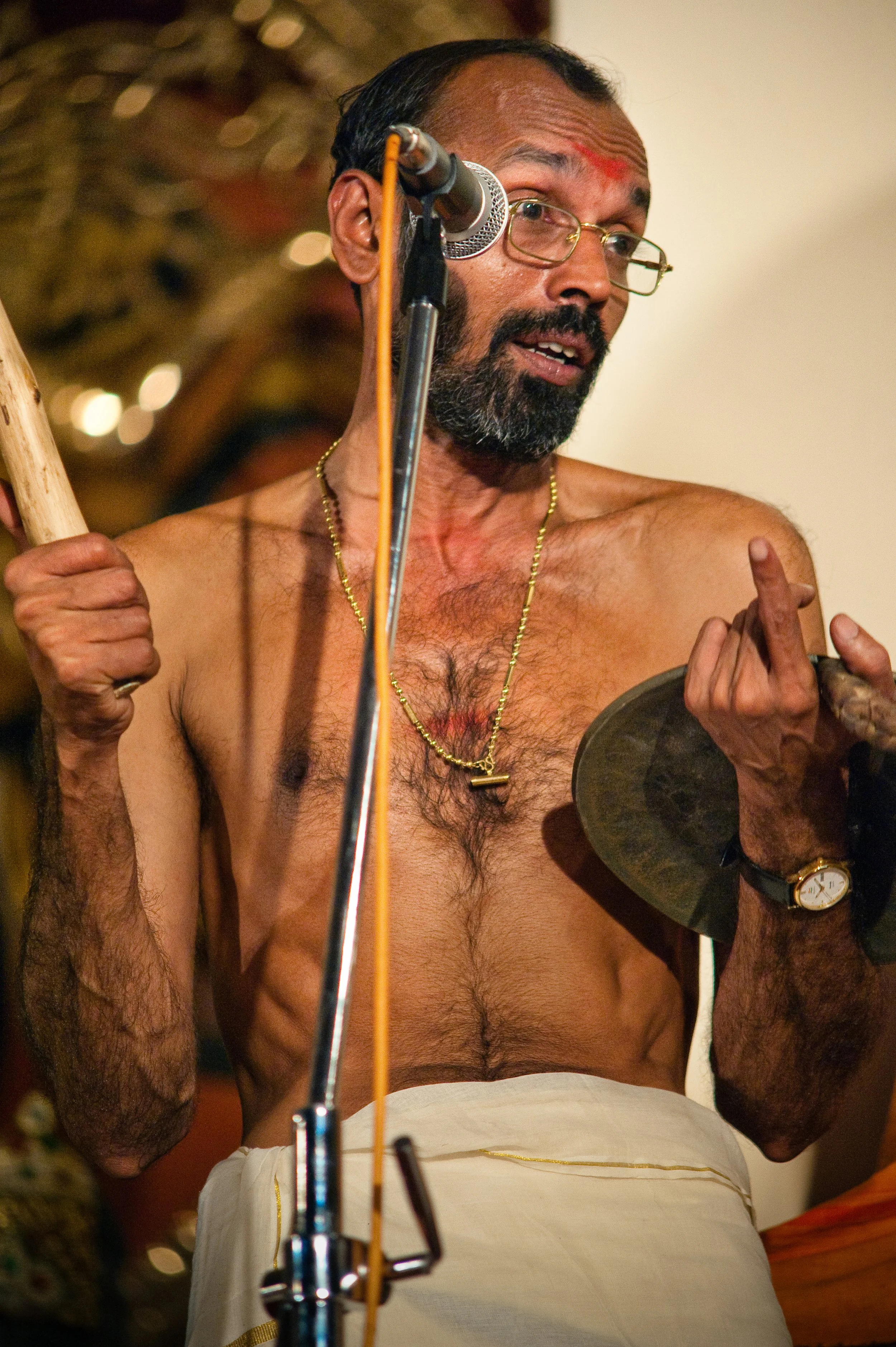 A shirtless man with glasses, a beard, and a gold necklace holding cymbals, standing in front of a microphone.