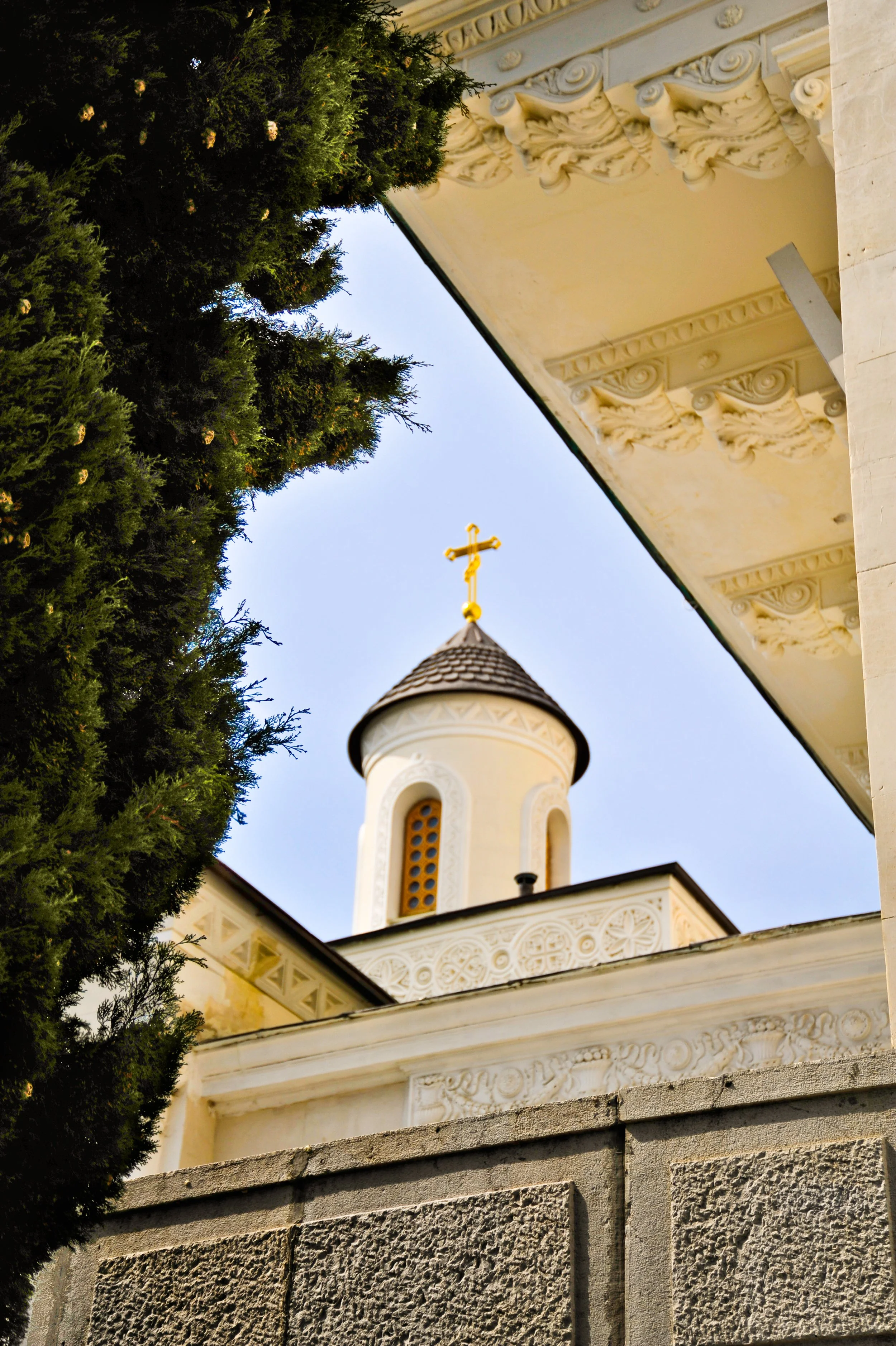 View of a church steeple with a cross on top, partially obscured by green trees and framed by ornate architectural details and a stone wall.