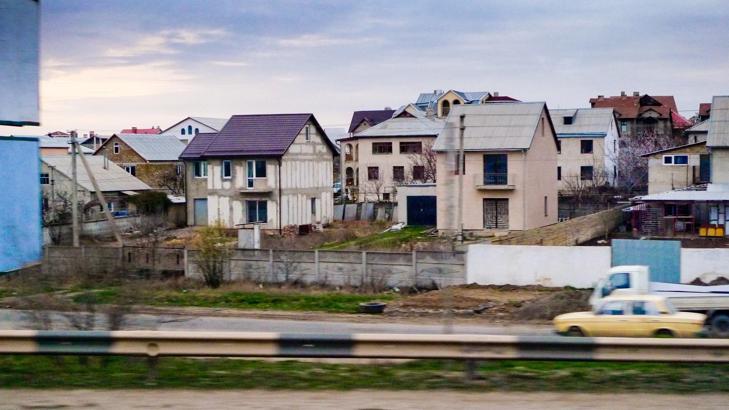 Residential area with houses under construction, a road with a moving yellow car, and a railroad track in the foreground.