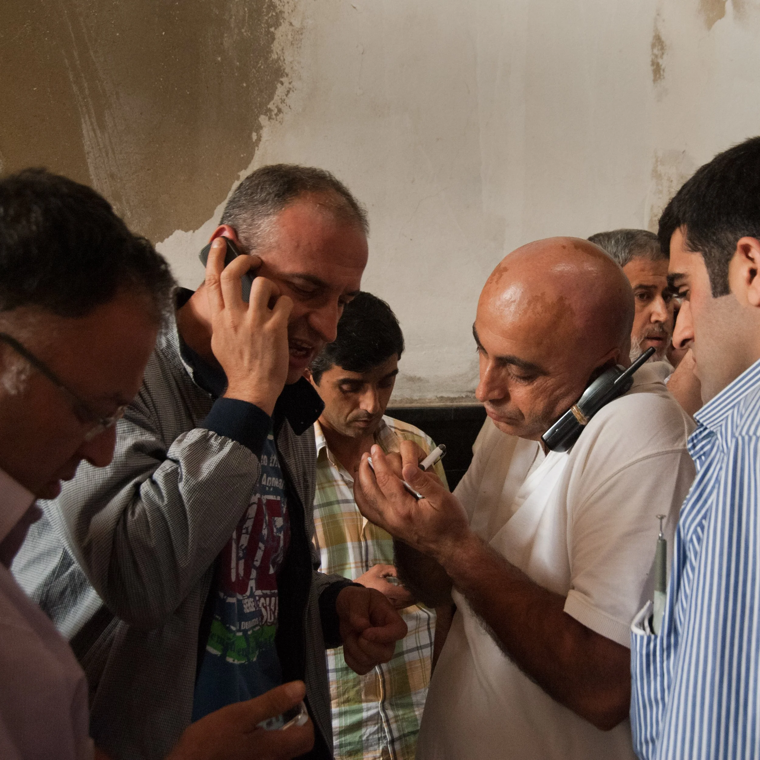 A group of men in a crowded room, talking on and holding phones, some looking at their phones.
