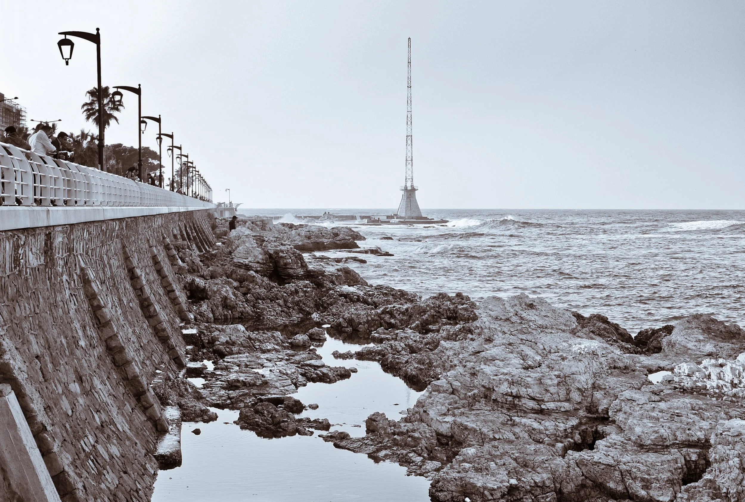 A seaside view with a stone seawall, street lamps, and people walking along the promenade. The rocky shoreline extends into the ocean, where waves are crashing. A tall lighthouse stands on a small platform in the water in the distance.