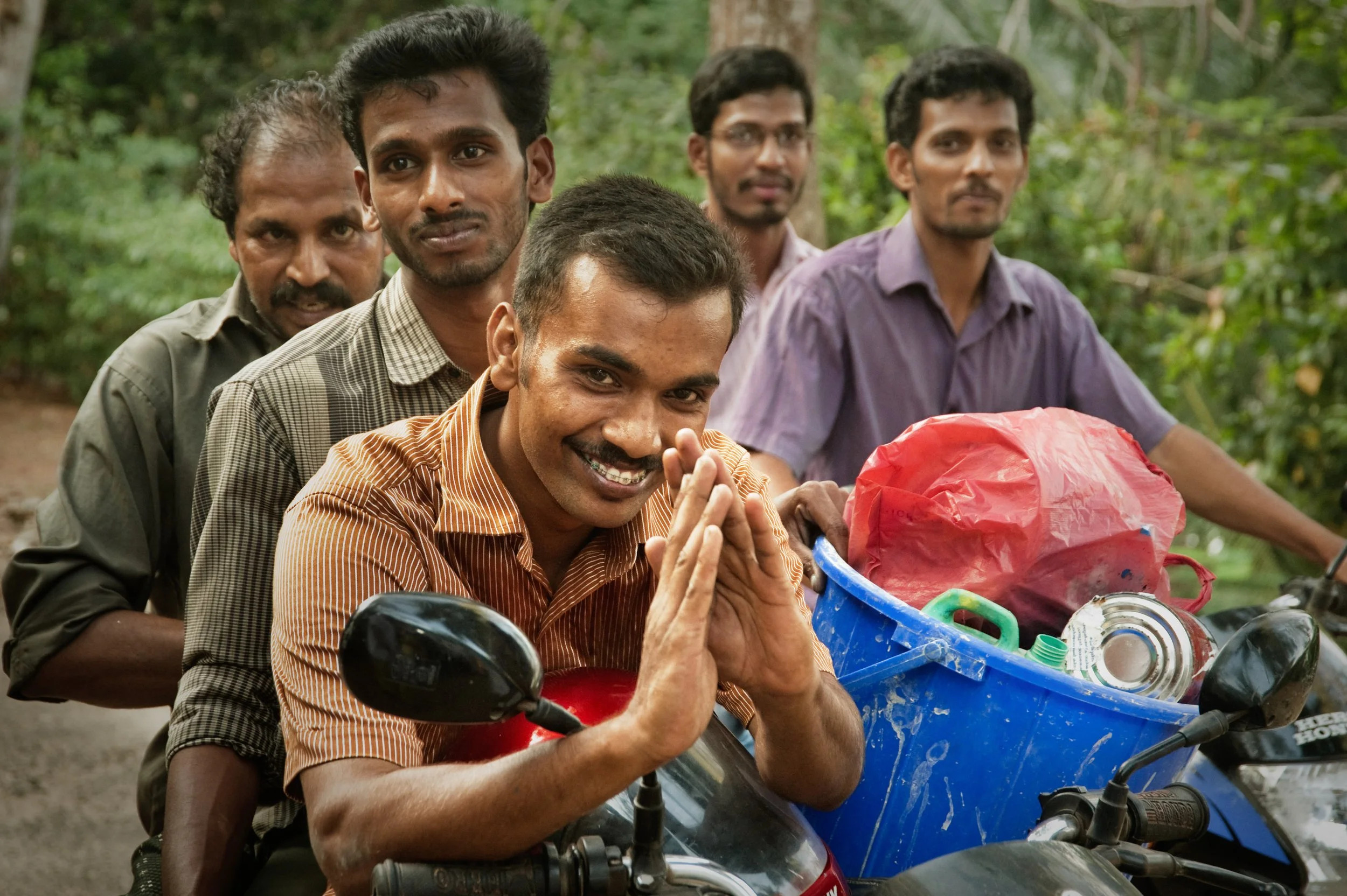 Five men on motorcycles with a blue cart filled with recyclables in a lush outdoor setting