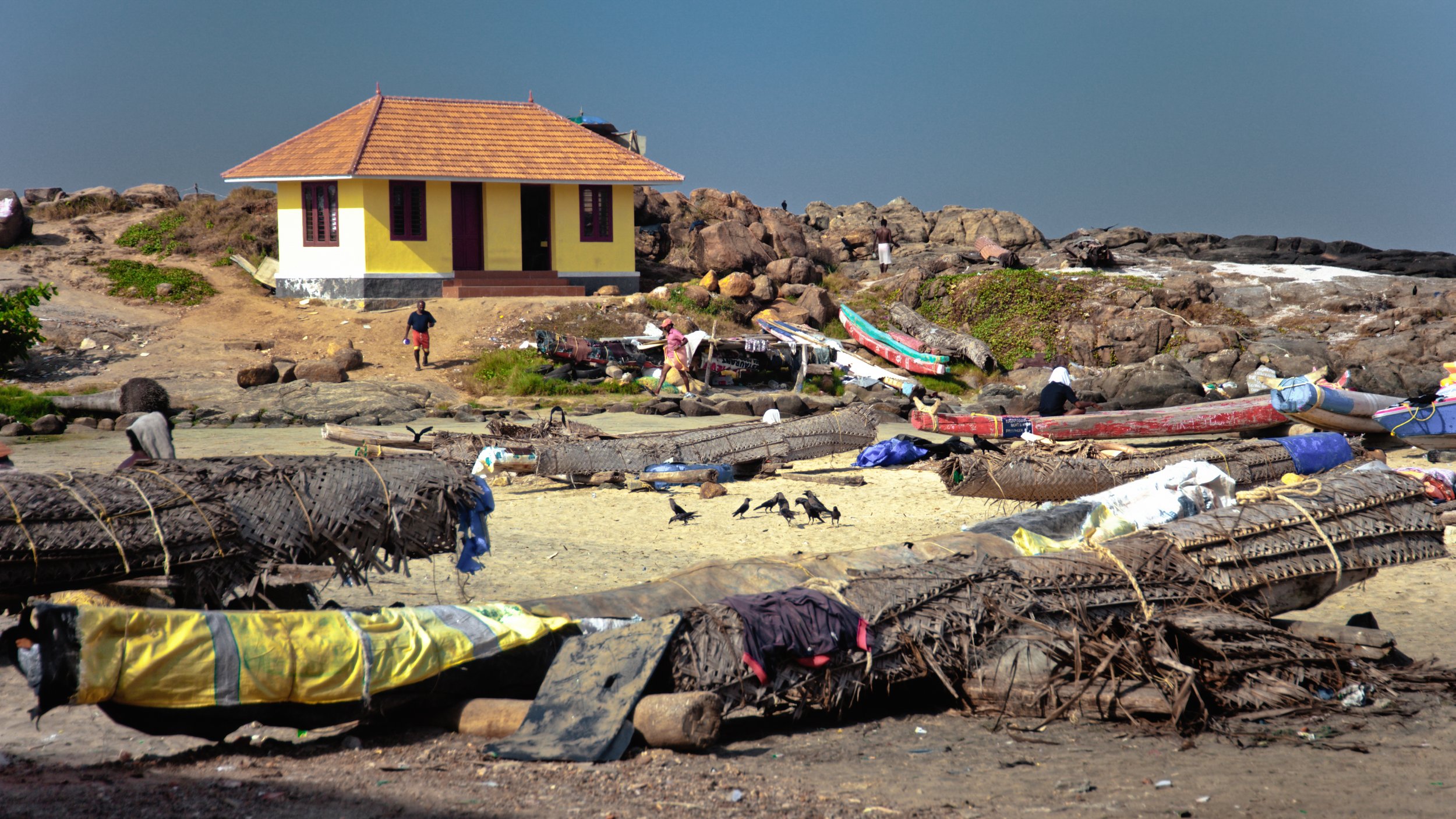 Beach scene with boats, people, and a yellow house on a rocky shoreline, with seagulls on the sand.