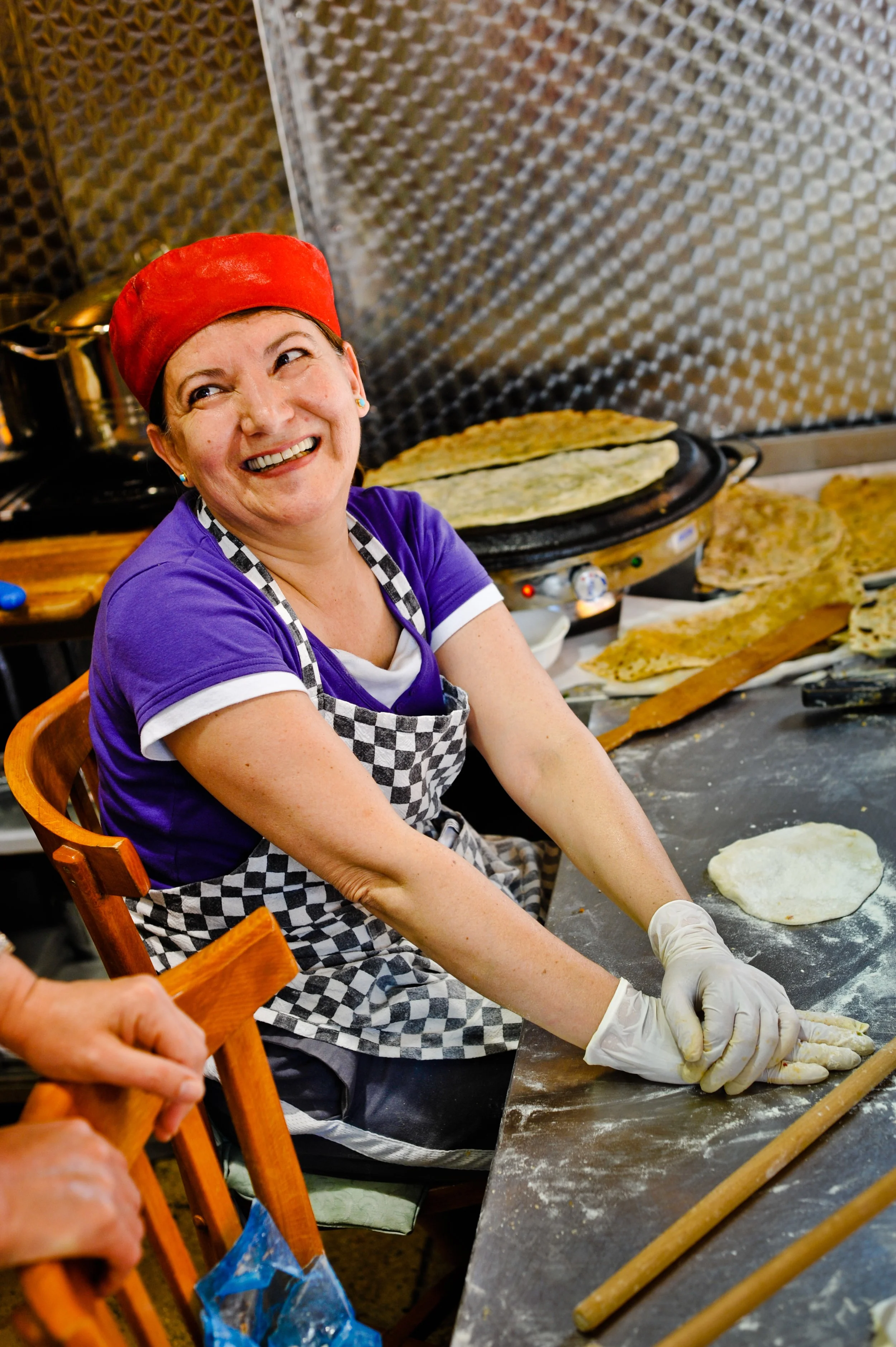 A woman with a red hat and purple shirt making pizza dough in a kitchen, smiling while kneading dough on a table.