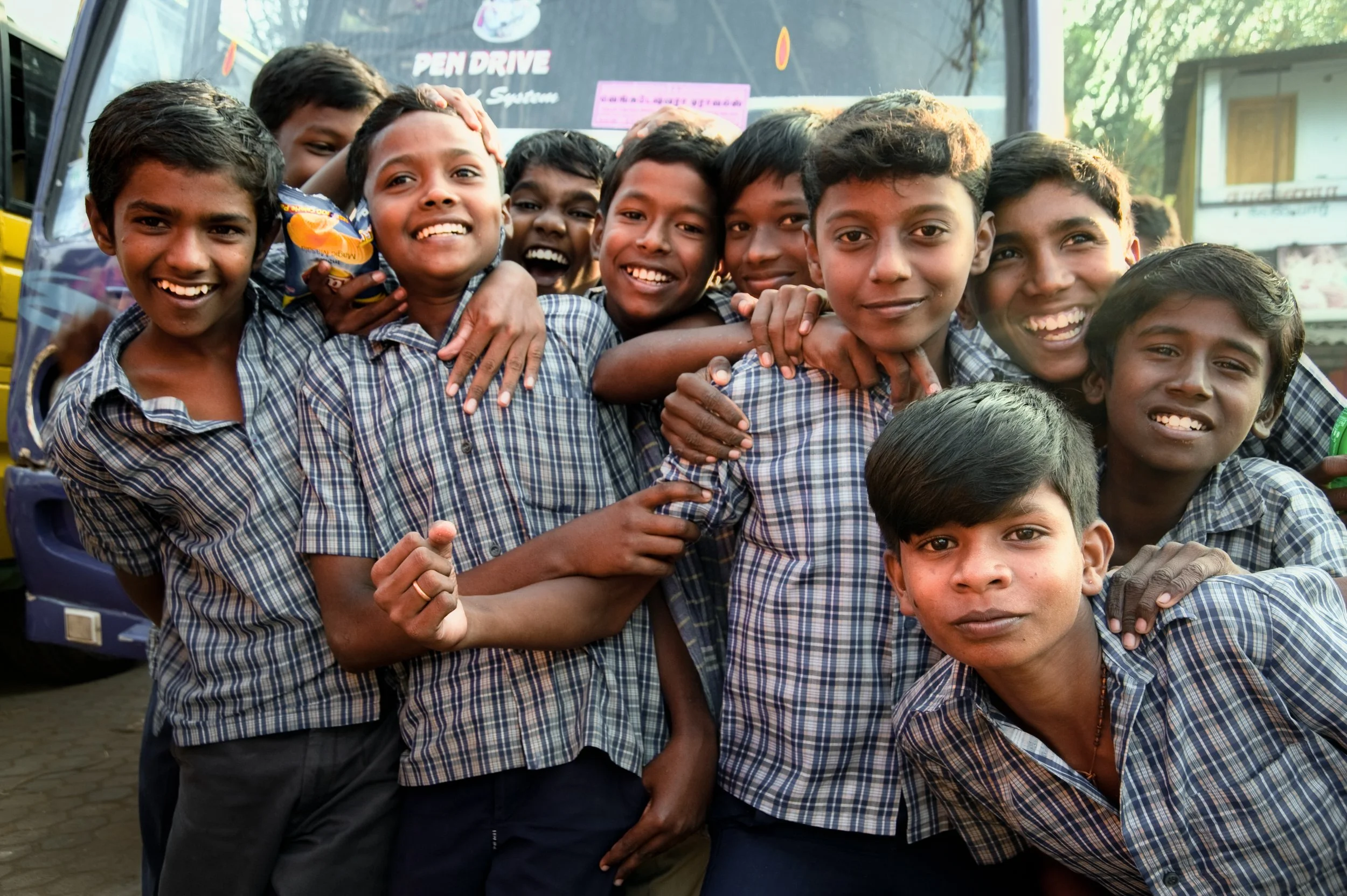 Group of smiling schoolboys wearing plaid uniforms, gathered together outdoors in front of a colorful vehicle.
