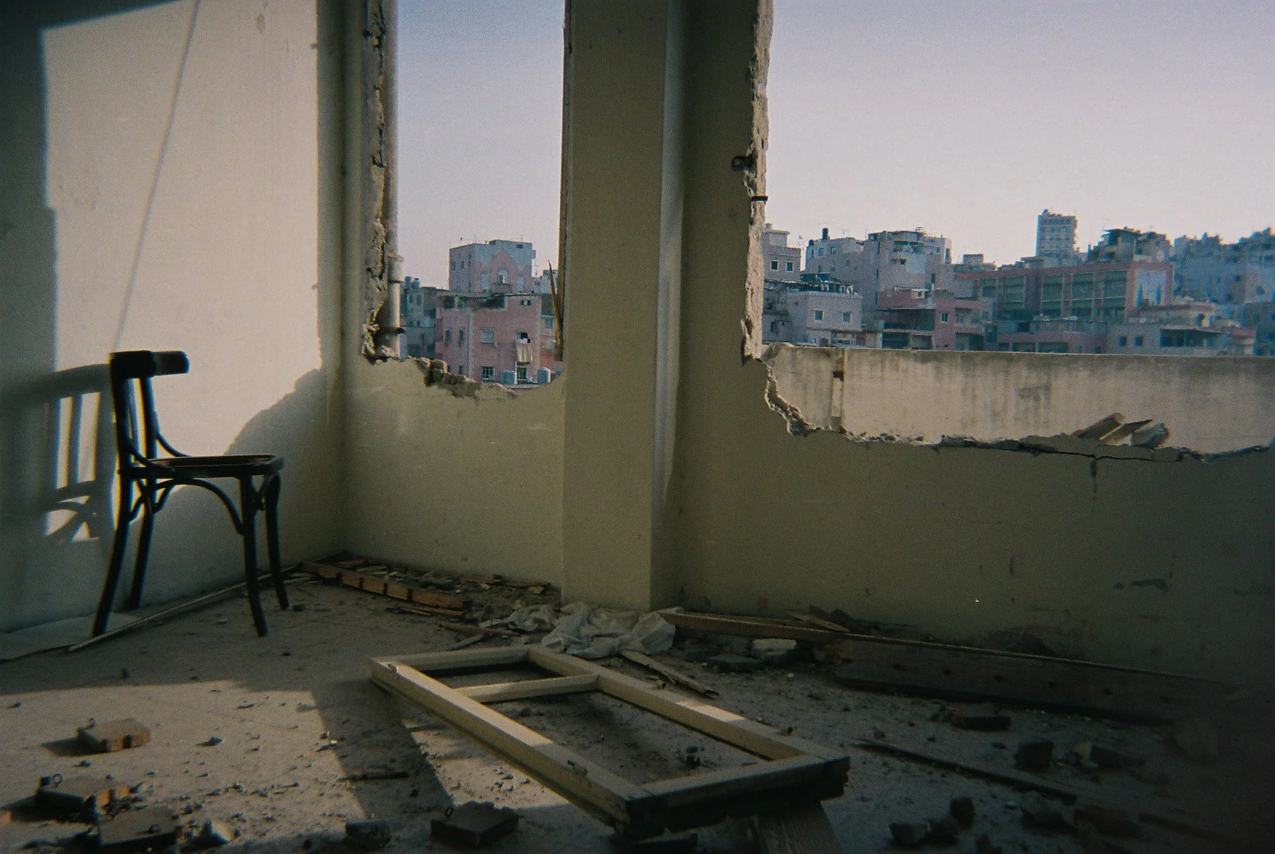 Interior of an unfinished building with broken walls and windows showing a cityscape of cluttered buildings, a black chair near the wall, and construction debris scattered on the floor.