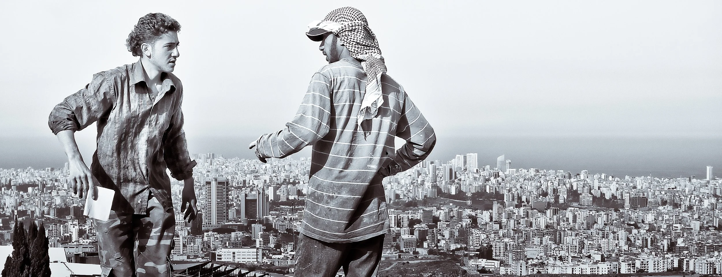 Two young men in casual clothing having a conversation on a hillside overlooking a city skyline.