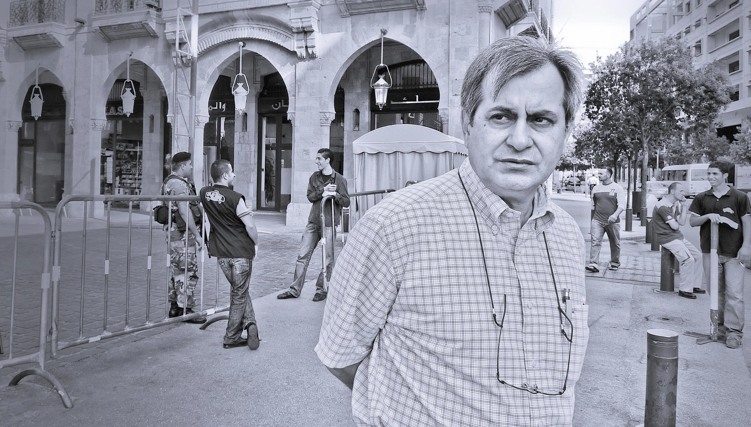 A man with glasses hanging around his neck standing on a city street with several young men in the background. Some are talking, some are sitting, and others are standing near barriers. The scene appears to be in an urban area with shops and trees.