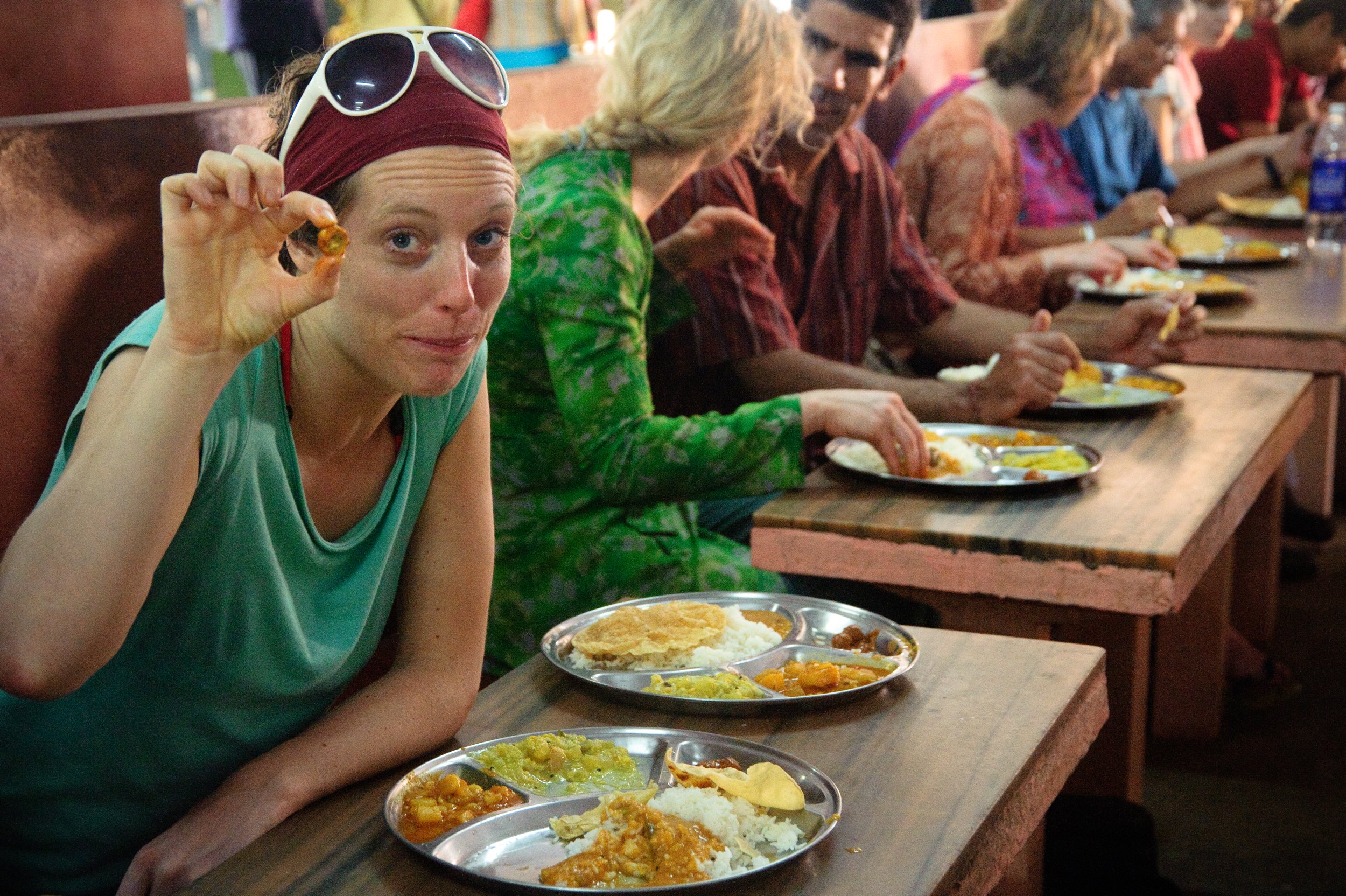 A woman in a green tank top and red headband holding a spice while sitting at a long wooden table with other people in a crowded dining area, with plates of rice, curry, and flatbread.