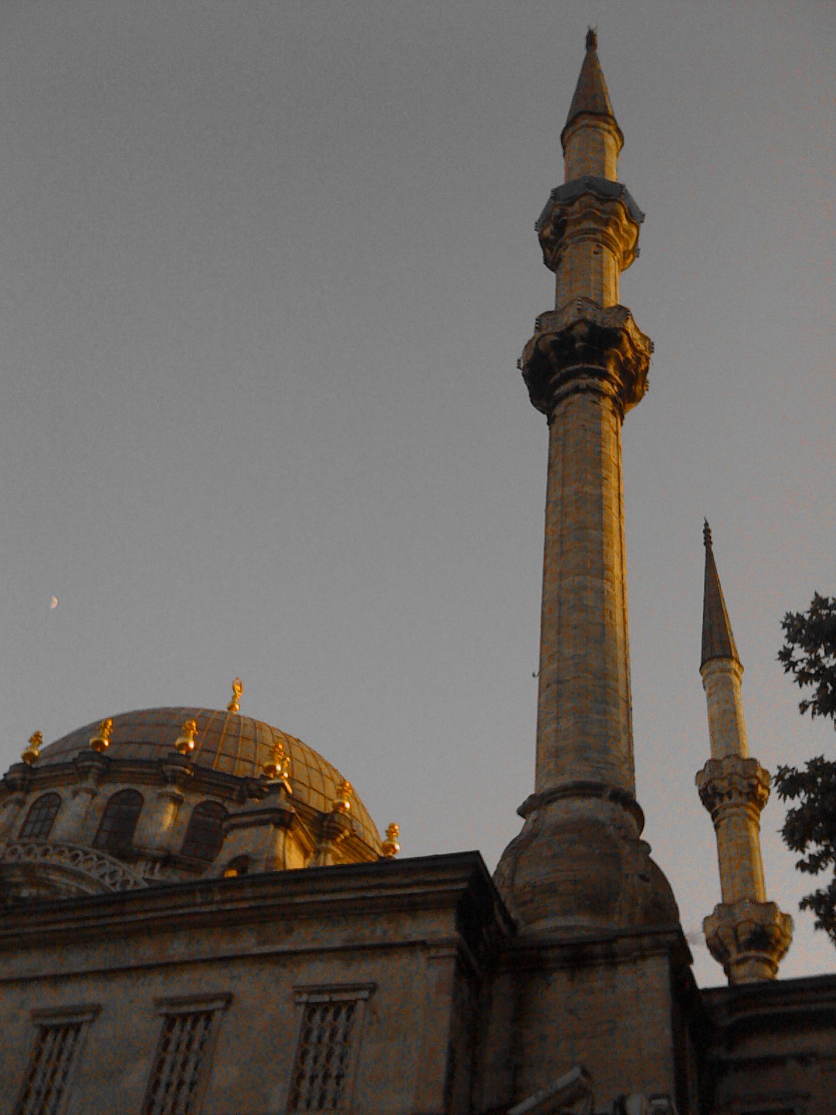 A mosque with tall minarets and a large dome, lit by warm evening light, against a darkening sky with a visible moon.
