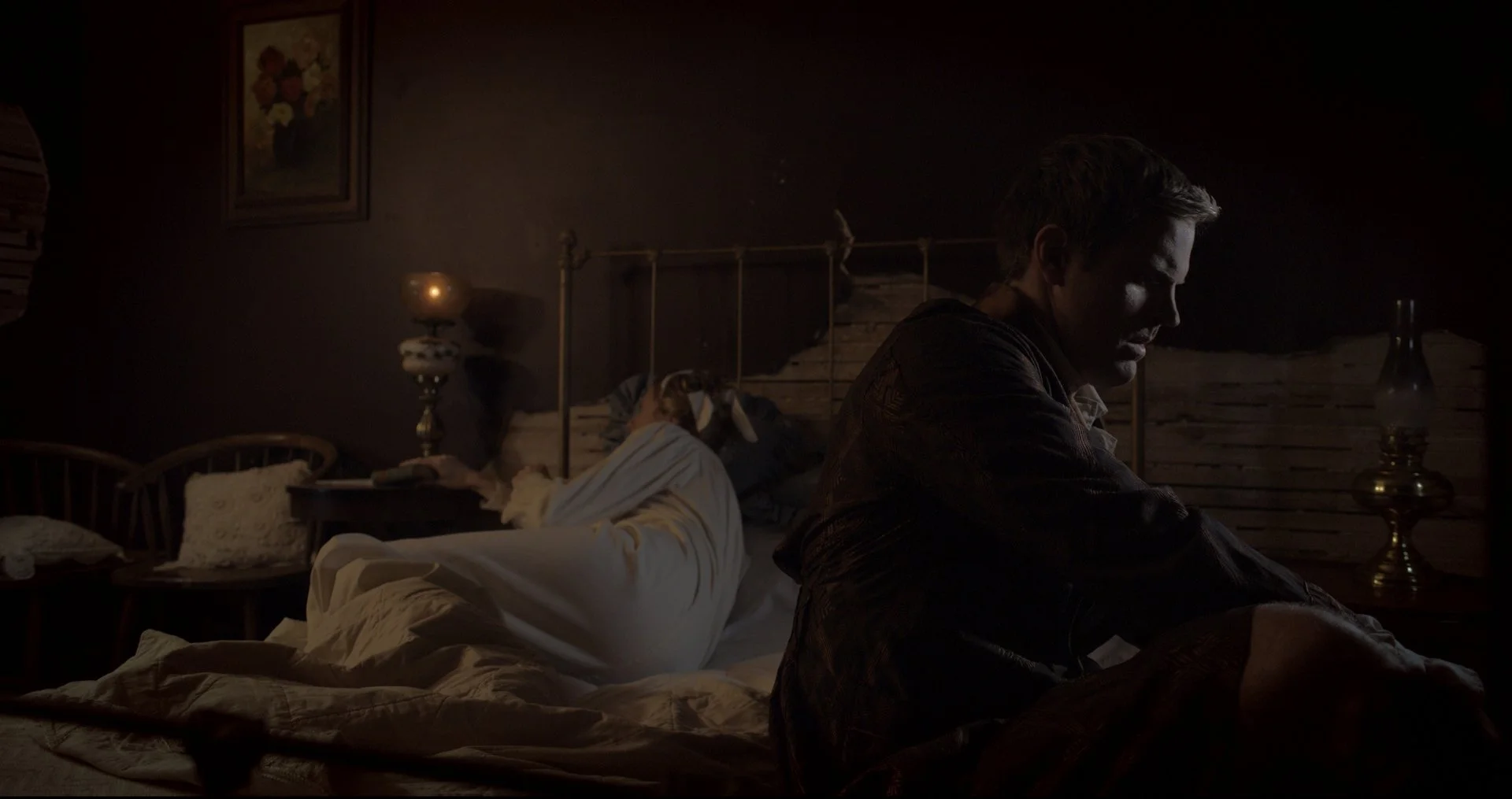 A dimly lit bedroom with a man sitting on the edge of the bed, facing away from the woman who is lying on the bed with her hair covering her face.