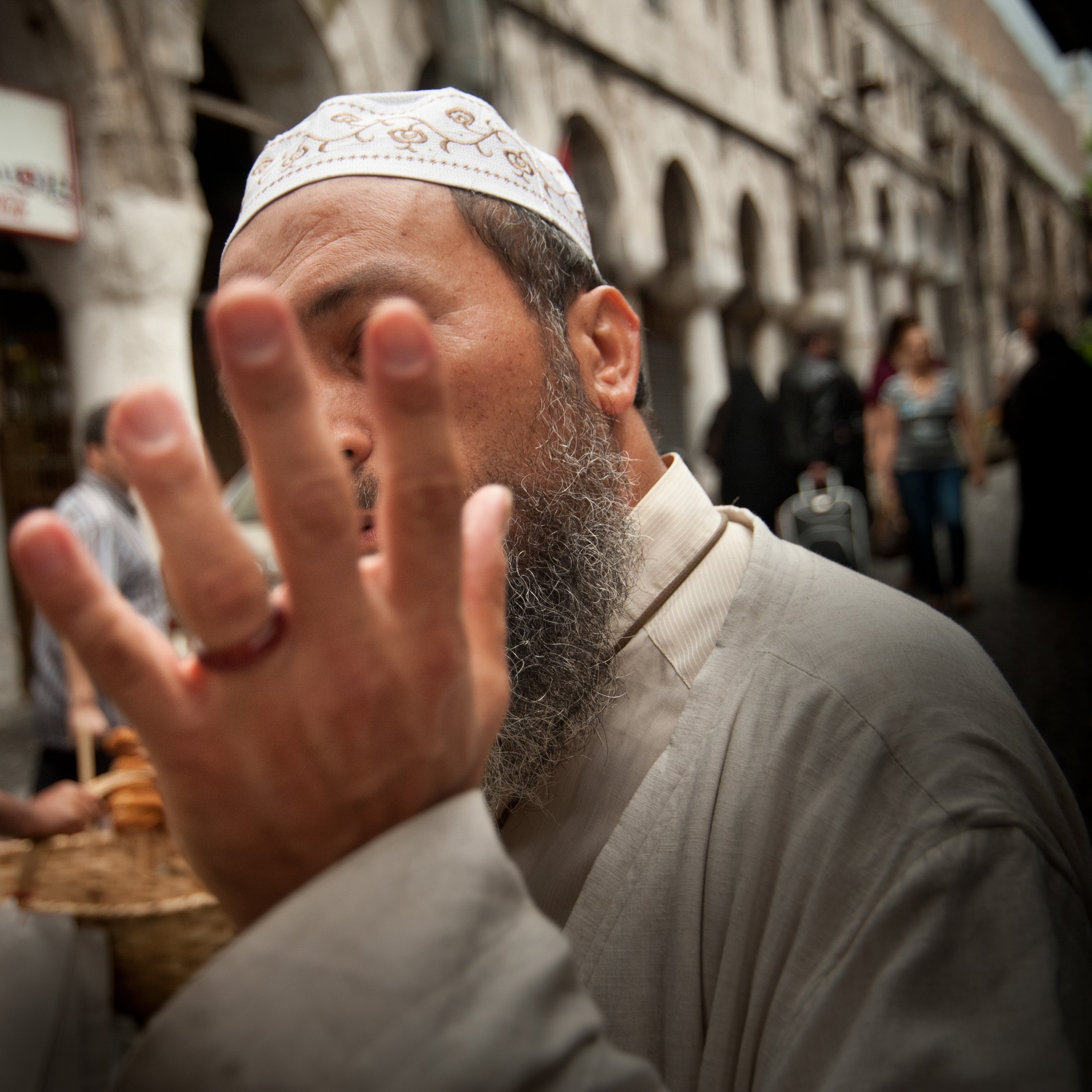 A man wearing traditional religious clothing and a white cap with gold embroidery, with a long beard, is raising his hand close to his face in a busy street scene. People and buildings are blurred in the background.