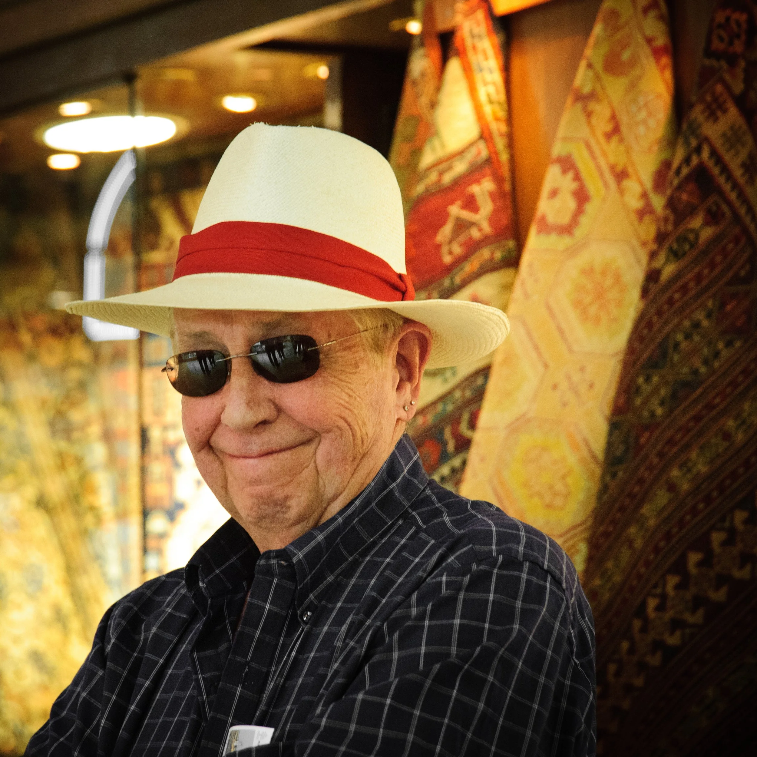 An elderly man smiling, wearing sunglasses, a wide-brimmed straw hat with a red band, and a plaid shirt, standing in front of colorful patterned rugs.