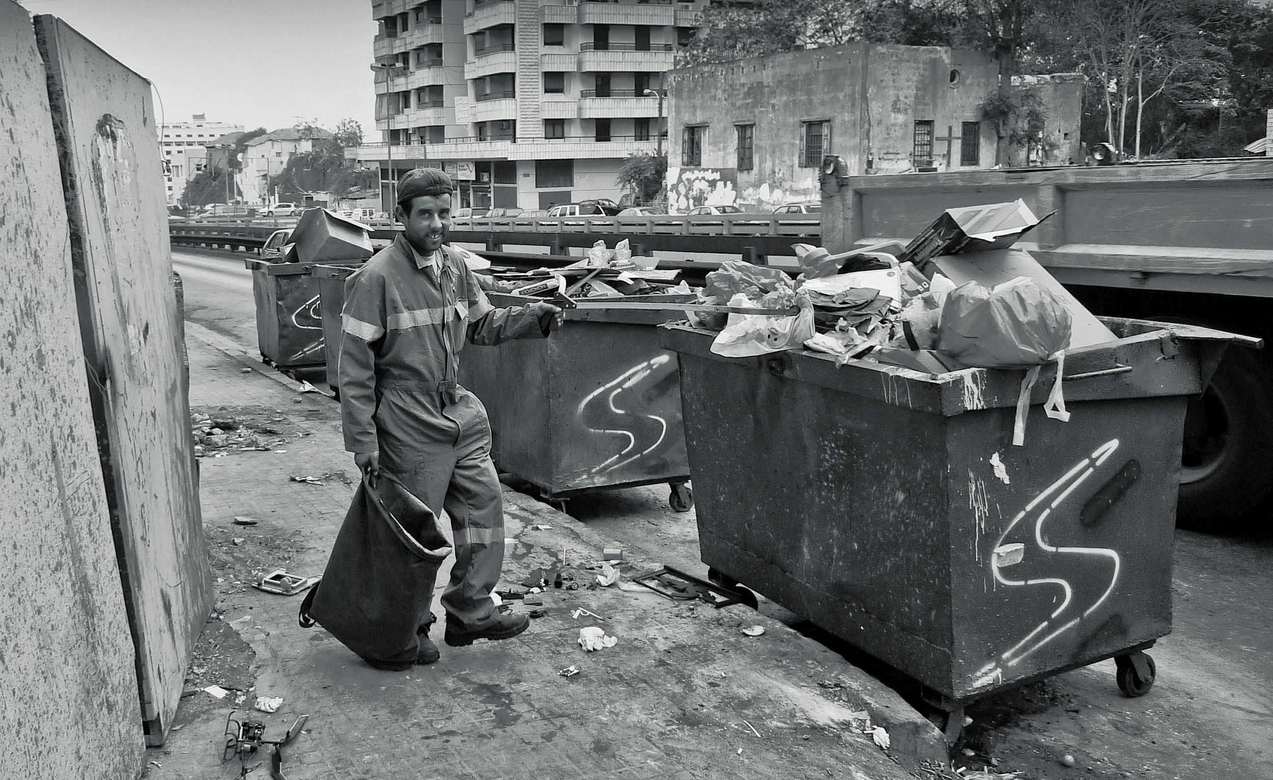 A man in work clothes smiling and holding a trash bag, standing next to overflowing garbage dumpsters with trash and debris on the street, in an urban area with buildings and a highway in the background.