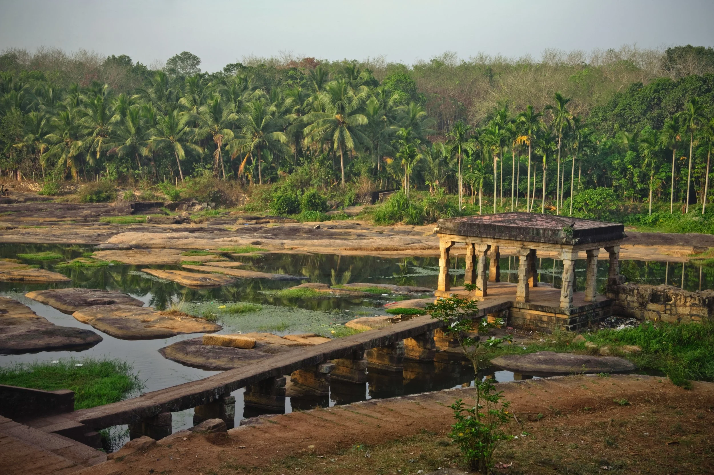Ancient stone temple with carved columns situated on a small hill overlooking a pond with stone walkways, surrounded by lush green trees and palms.
